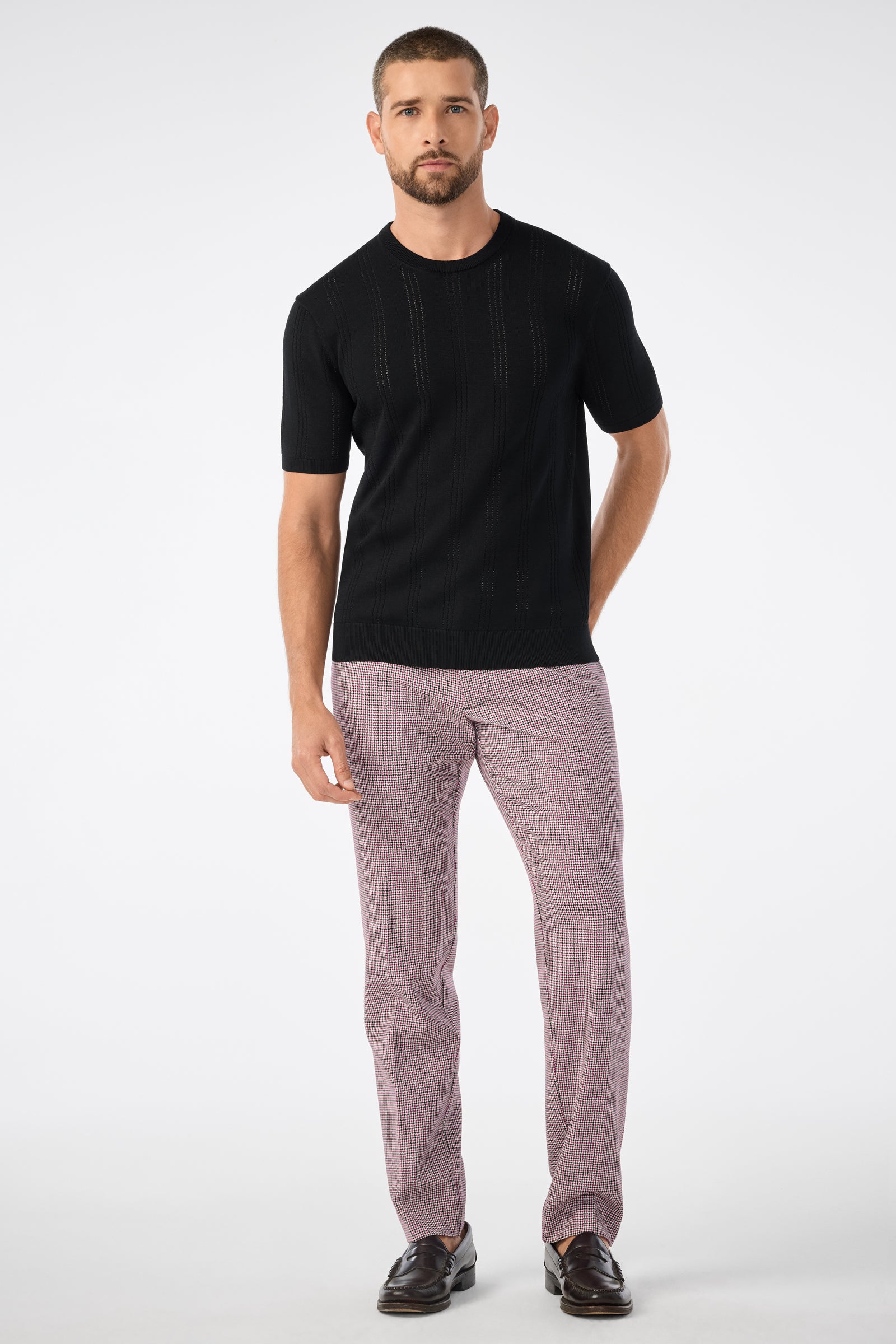 A man with short hair and a beard stands against a white background, wearing the Carlo Ss Crewneck black short-sleeve sweater, pink and gray checkered pants, and brown loafers, with one hand in his pocket.