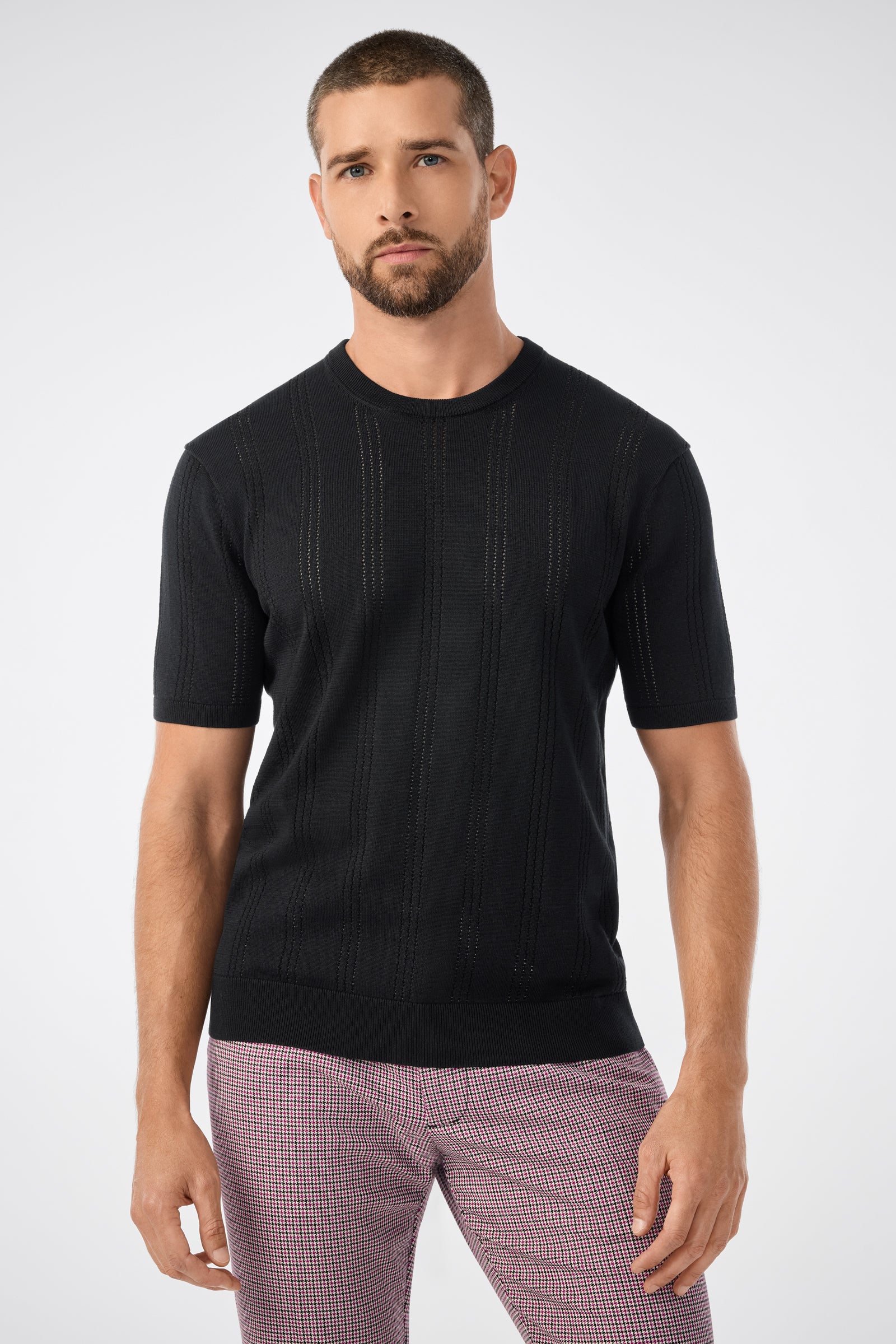 A man with short hair and a beard wears the black Carlo Ss Crewneck sweater with subtle vertical stripes and pink checkered pants, standing against a plain light gray background.