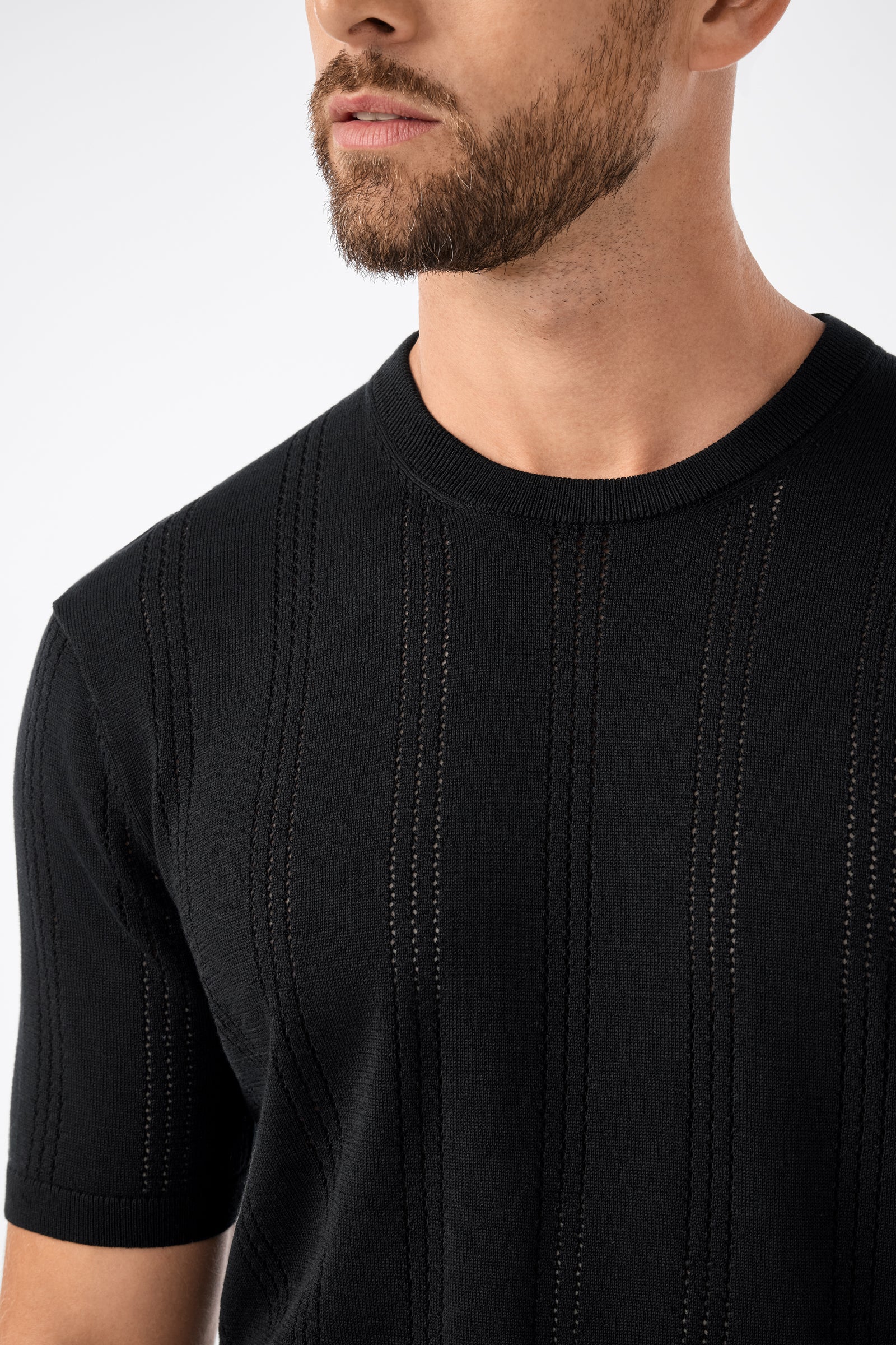 A man with a short beard wears the black Carlo Ss Crewneck, featuring vertical textured and openwork stripes. The image is cropped to highlight his upper torso, neck, and part of his face.