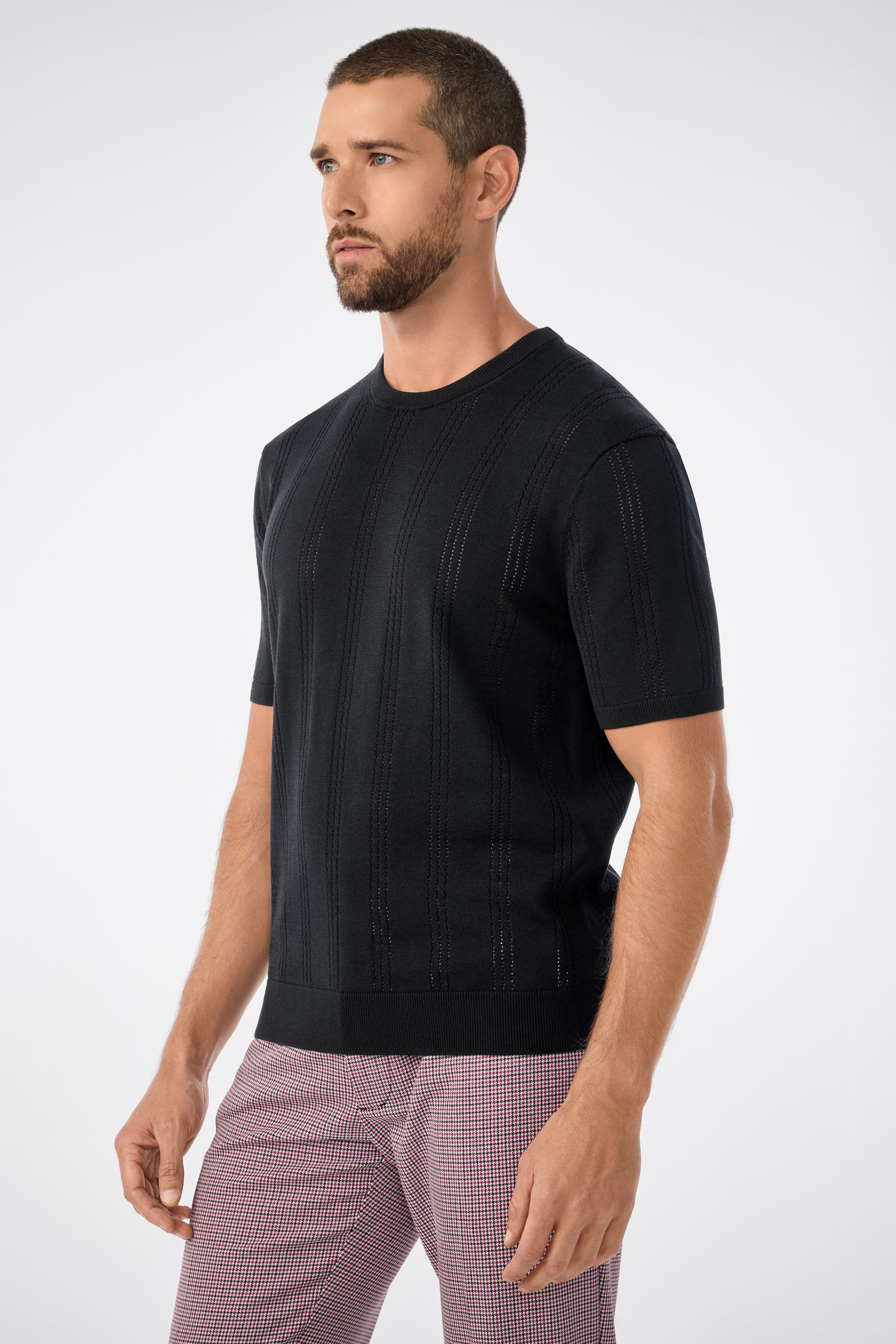 A man with short hair and a beard wears the black Carlo Ss Crewneck sweater and light plaid pants, standing against a plain light background.