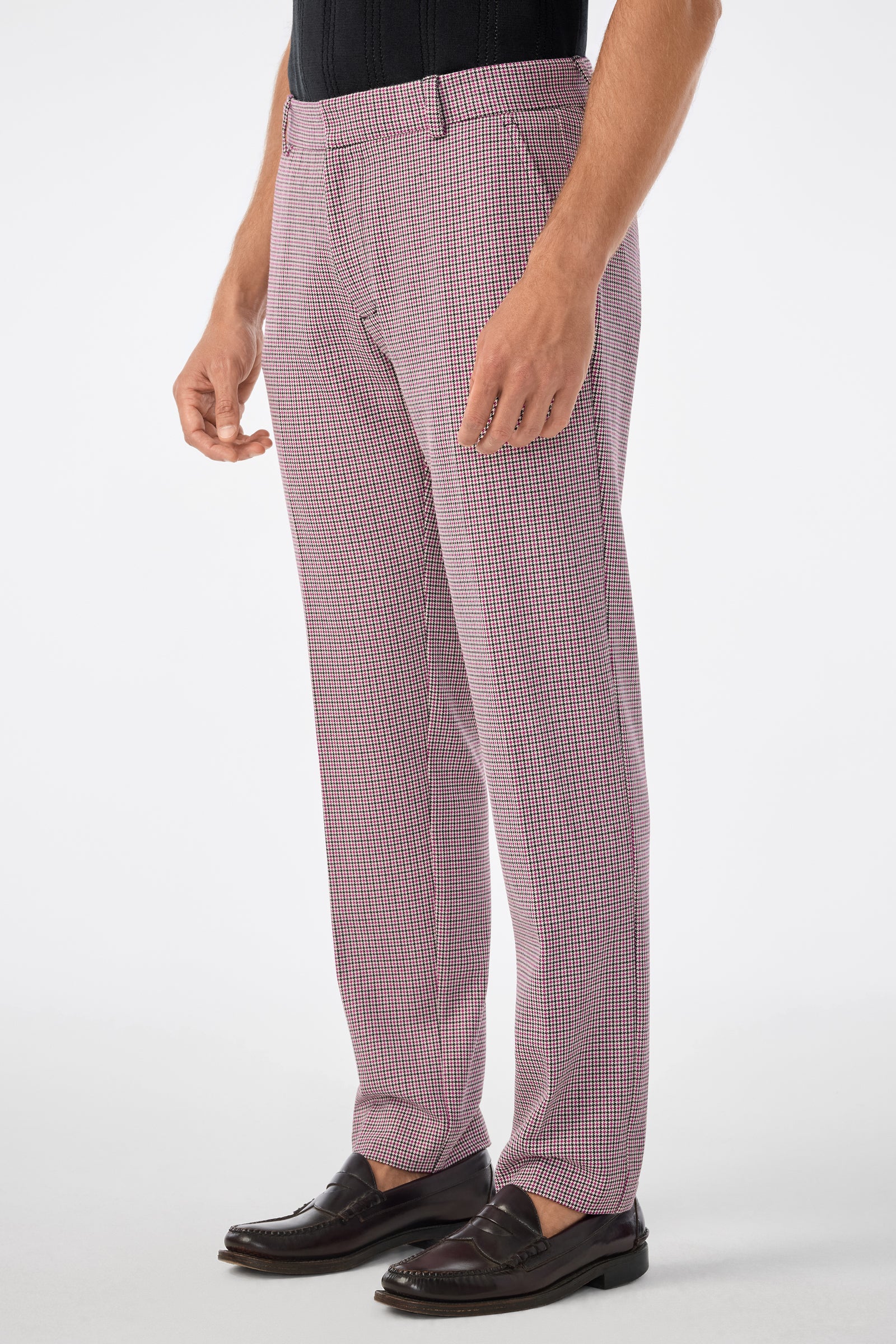 A person wearing the Clyde Slim Trouser in a pink and white checkered pattern, paired with a black tucked-in shirt and black loafers, standing against a plain light background.