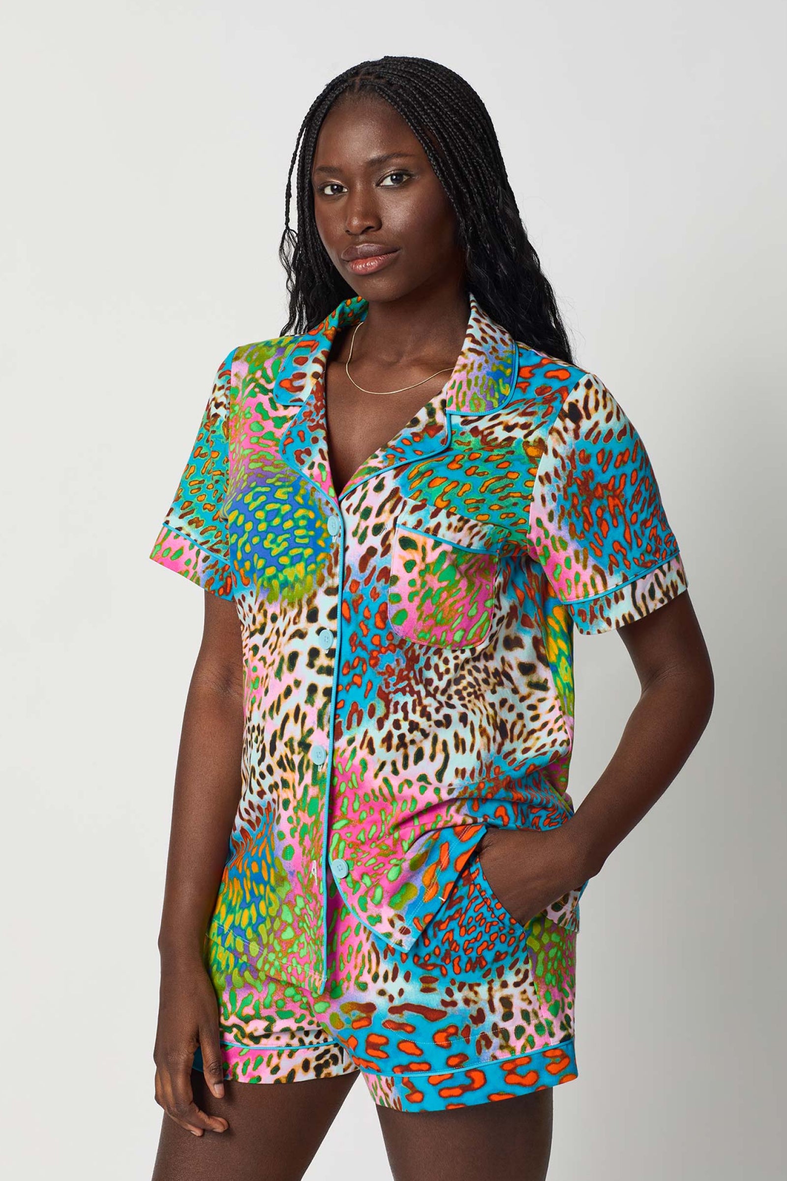 A woman with long braided hair wears the Shorty PJ Set - Spray Leopard, a colorful, short-sleeved shorts set with a vibrant blue, green, pink, and orange animal print pattern, standing against a plain light background.