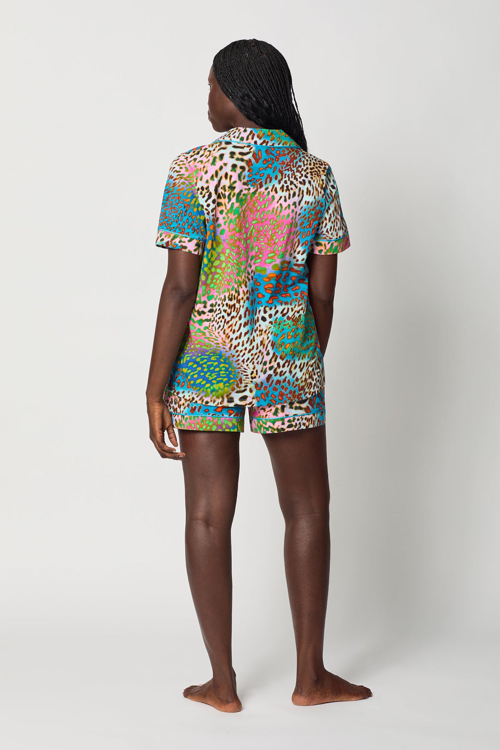 A person with dark braided hair stands barefoot, facing away from the camera, wearing the Shorty PJ Set - Spray Leopard: a colorful, short-sleeved, collared shirt and matching shorts with a vibrant leopard print.