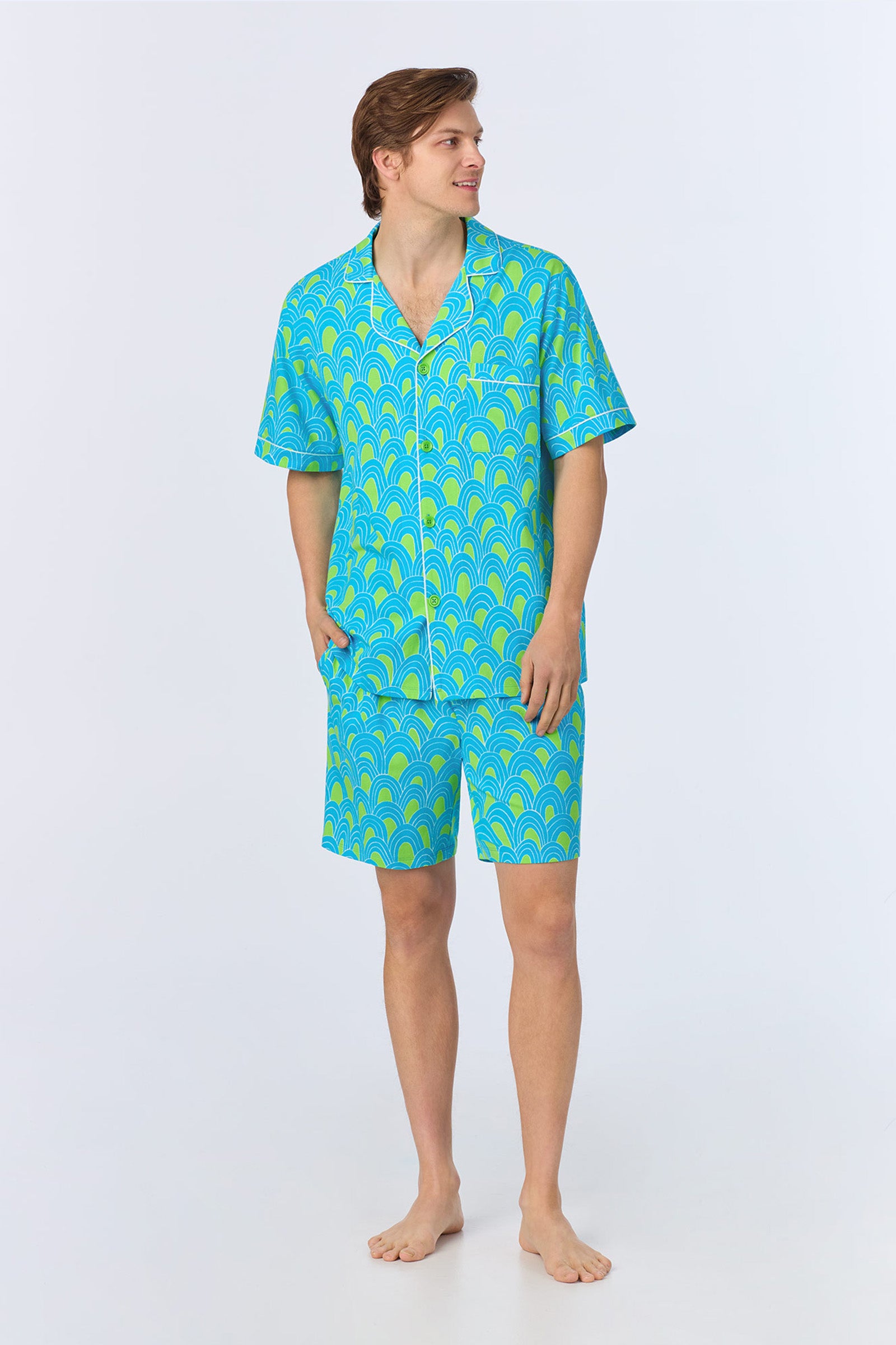 A person stands barefoot against a plain light background, wearing the Mens Boxer PJ Set - Geo Stretch Jersey: a matching short-sleeve button-up shirt and shorts in a blue and green scallop pattern, looking to the side.