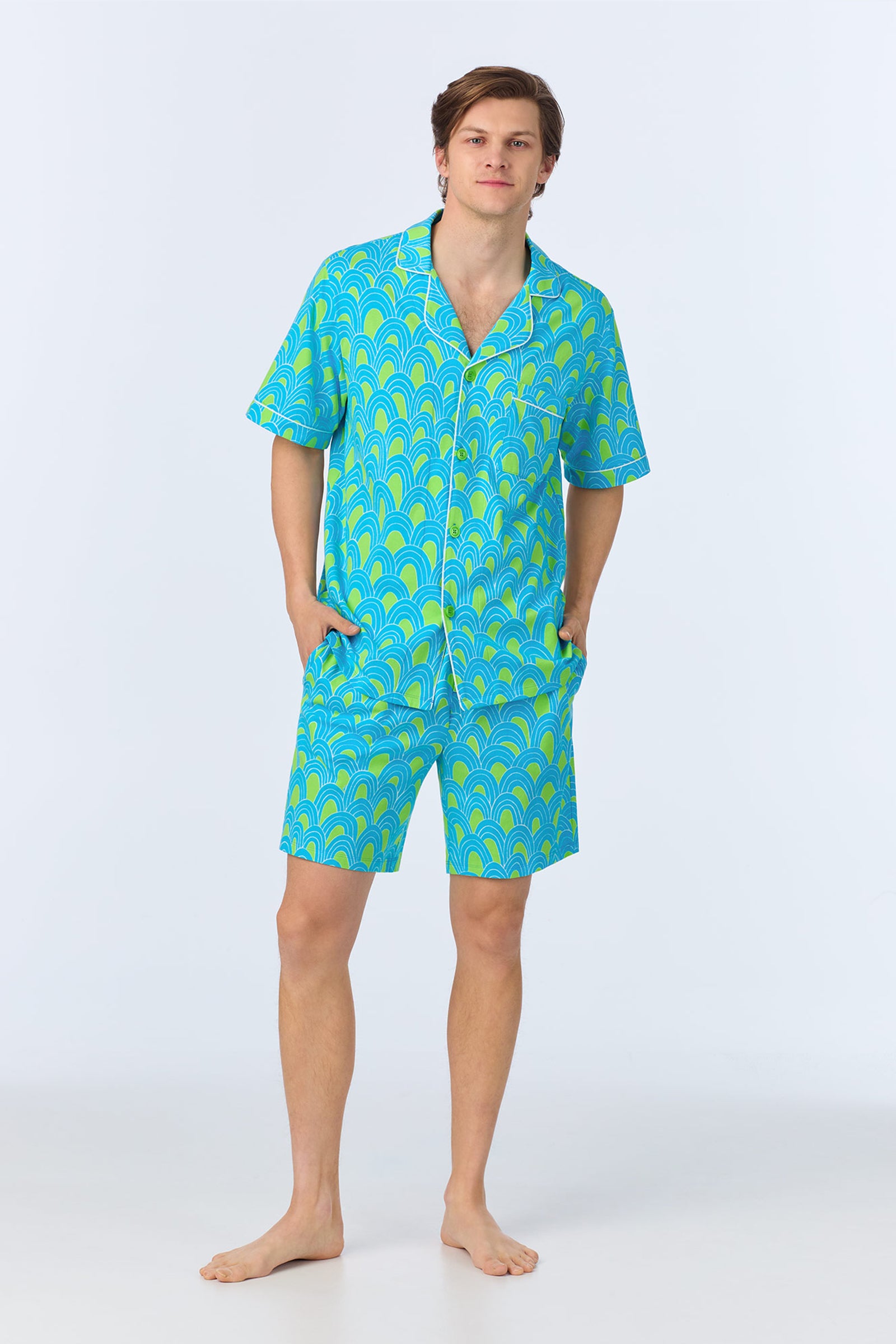 A person stands barefoot wearing the Mens Boxer PJ Set - Geo Stretch Jersey in turquoise, featuring a matching short-sleeve button-up shirt and shorts with a geometric pattern, posing hands in pockets against a plain light background.