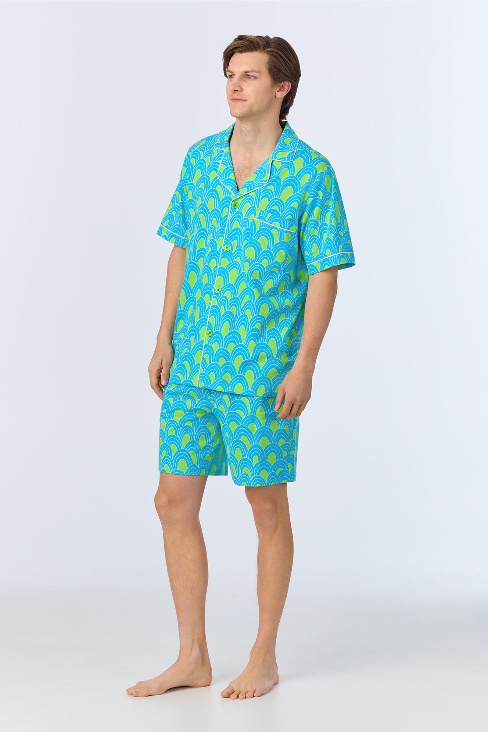 A man stands barefoot in the Mens Boxer PJ Set - Geo Stretch Jersey, featuring matching blue and green fan-patterned short-sleeve button-up shirt and shorts, against a plain light background.