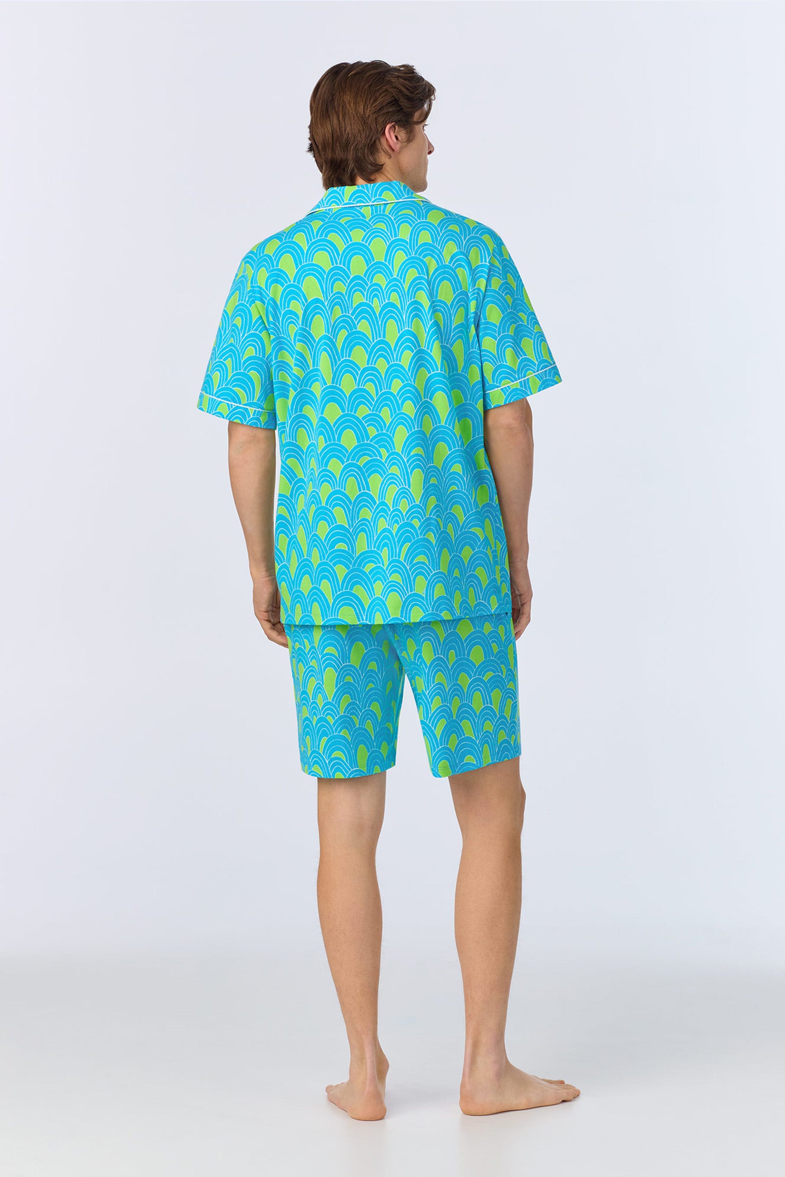 A barefoot man faces away on a light surface, wearing the Mens Boxer PJ Set - Geo Stretch Jersey, which features a matching short-sleeve shirt and shorts with a bright blue and green geometric pattern. The background is plain white.