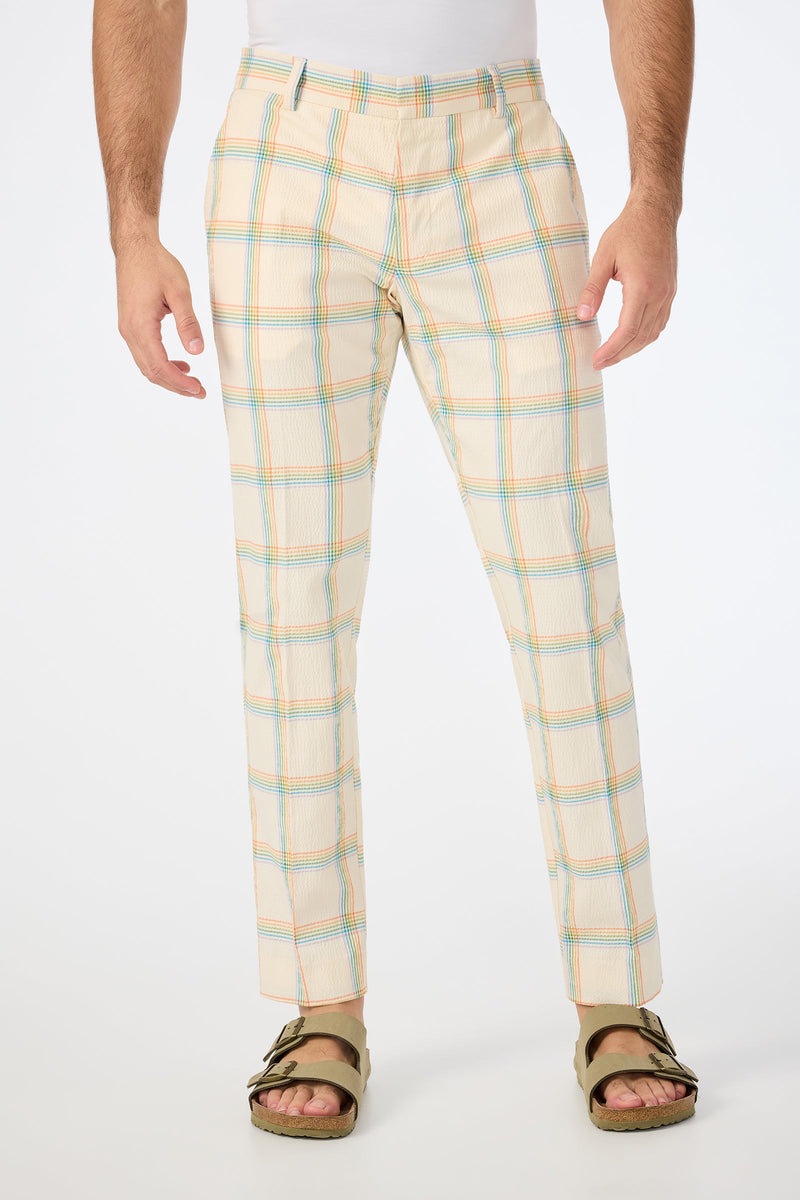 A person wears the Clyde Slim Trouser in Pride Plaid—cream with light blue, orange, and green stripes—paired with green sandals and a white shirt, standing against a plain white background.