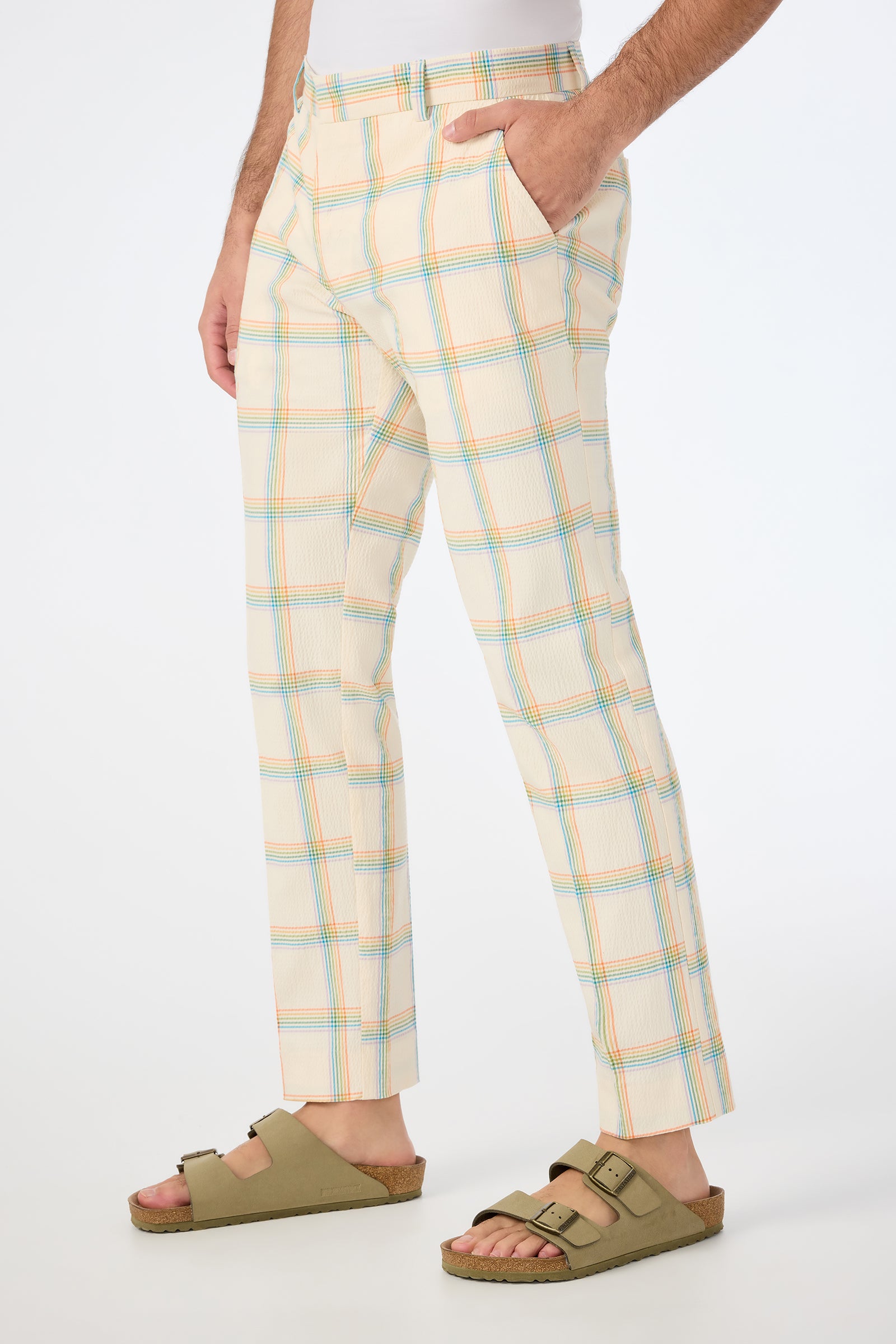 A person is shown in the Clyde Slim Trouser - Pride Plaid, featuring a beige plaid with blue, orange, and yellow lines, standing sideways with hands in pockets and wearing tan Birkenstock sandals against a white background.