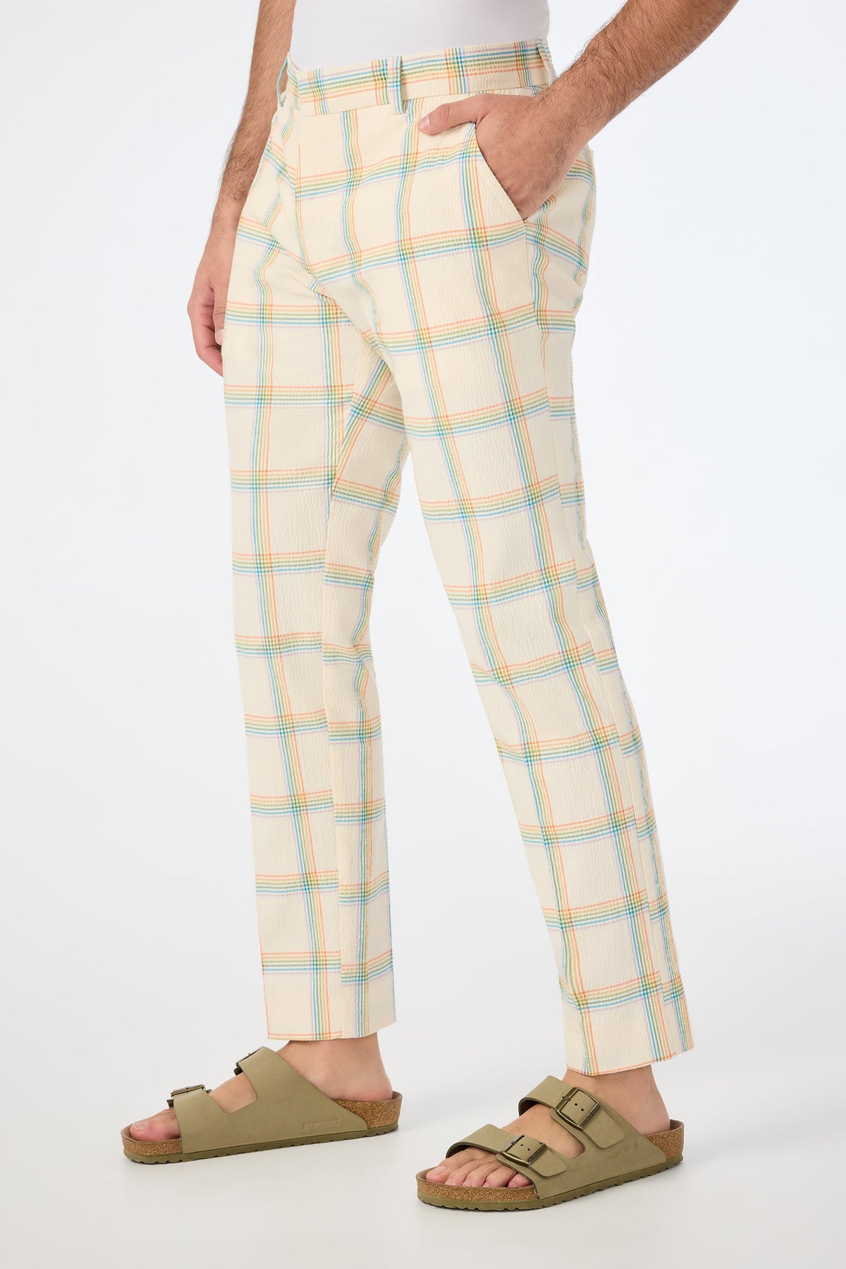 A person wears the Clyde Slim Trouser in Pride Plaid—a cream-colored pant with a pastel plaid pattern—hands in pockets, paired with light brown sandals, against a plain white background.