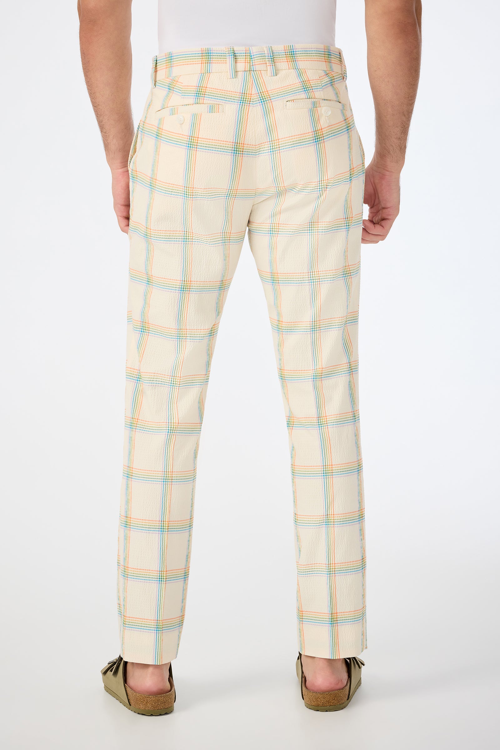 Someone wearing the Clyde Slim Trouser - Pride Plaid, featuring a blue, yellow, and orange plaid pattern with buttoned back pockets. Shown from the back, paired with tan moccasins and a white shirt.