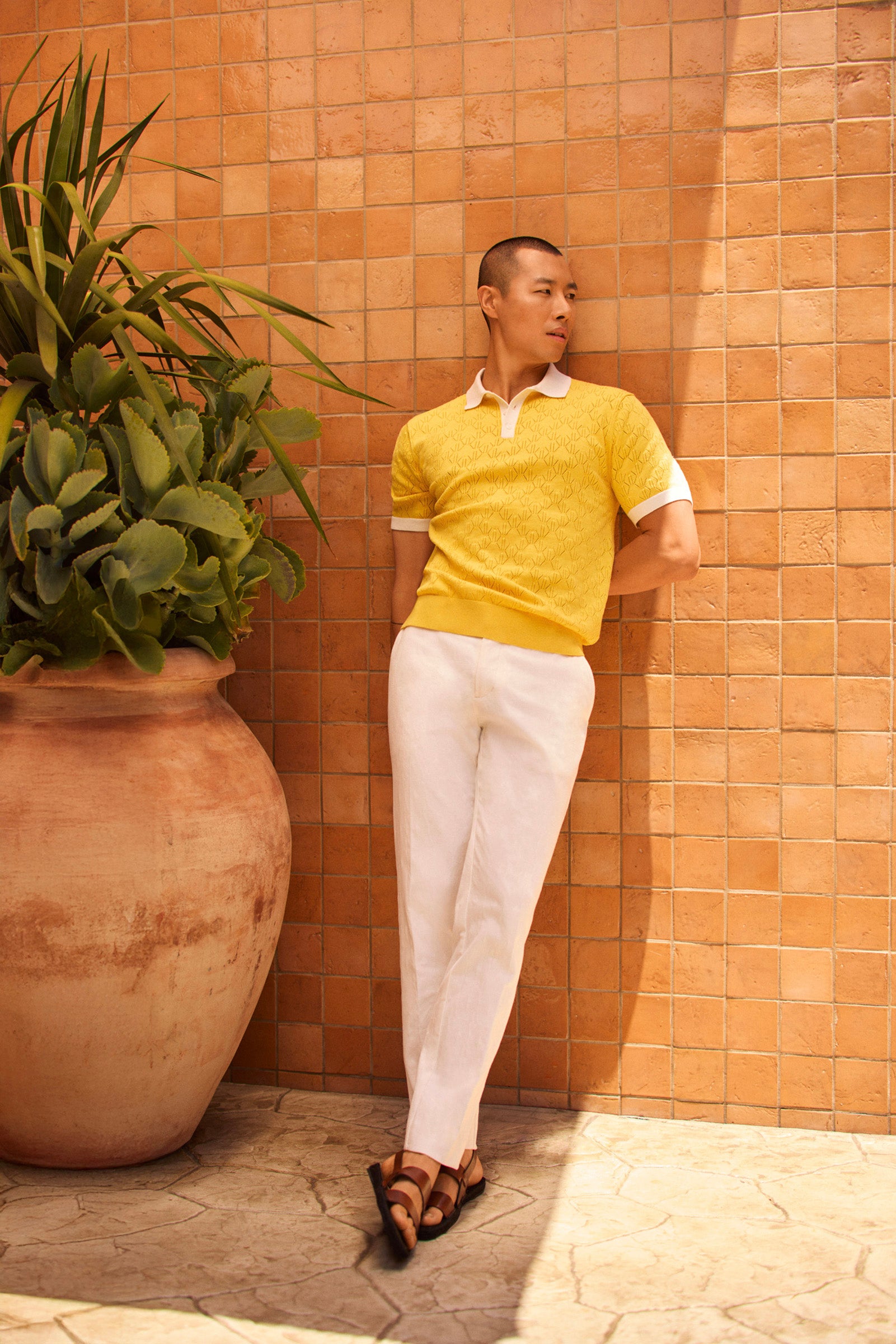 A person stands by a peach-tiled wall near a potted plant, wearing the yellow Luca Short-Sleeve Polo over a white collared shirt, paired with white pants and brown sandals.