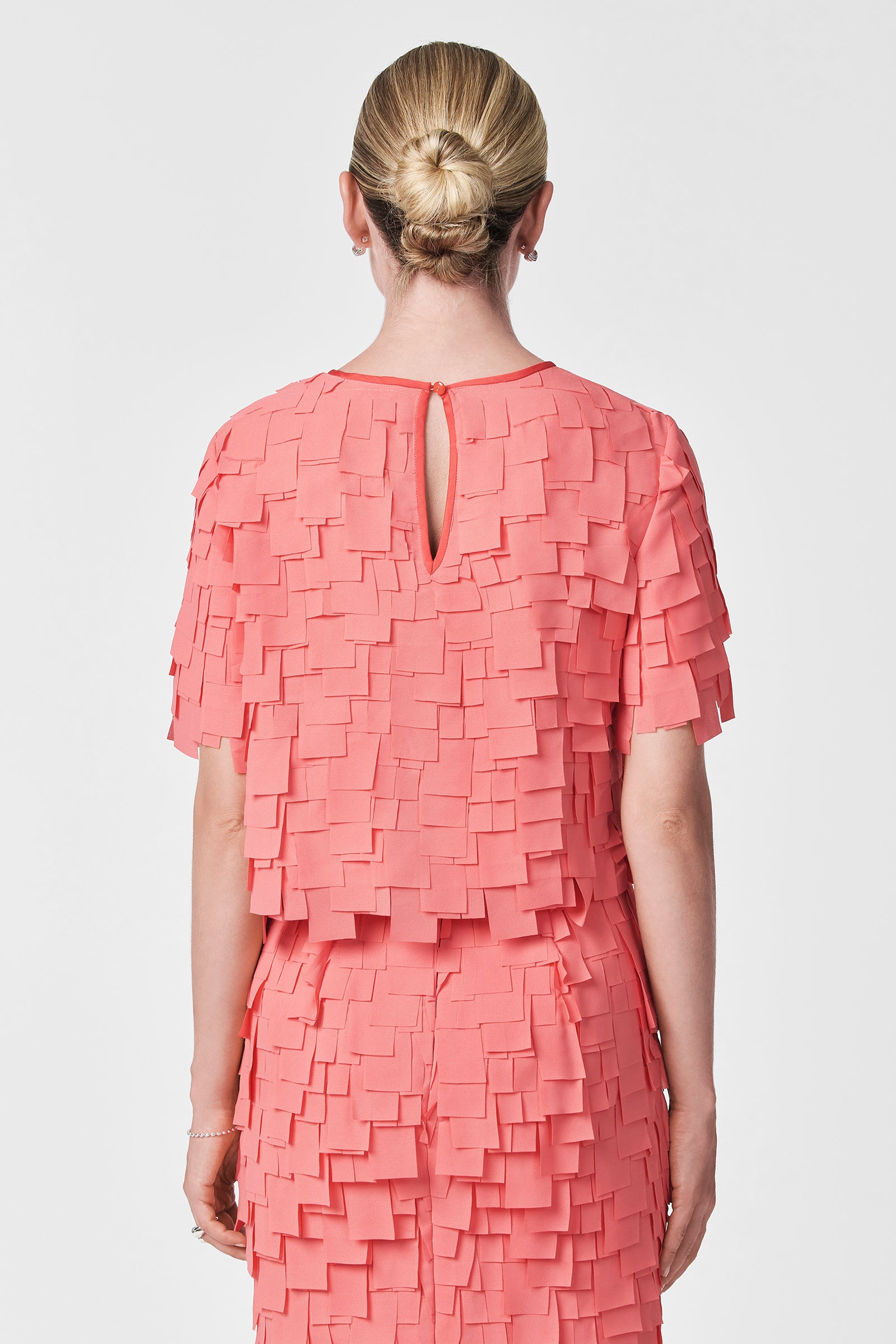 A person with blonde hair in a bun is seen from the back against a white background, wearing the Serilda Short-Sleeve Top - Flutter Chiffon in textured coral pink with layered square chiffon pieces.