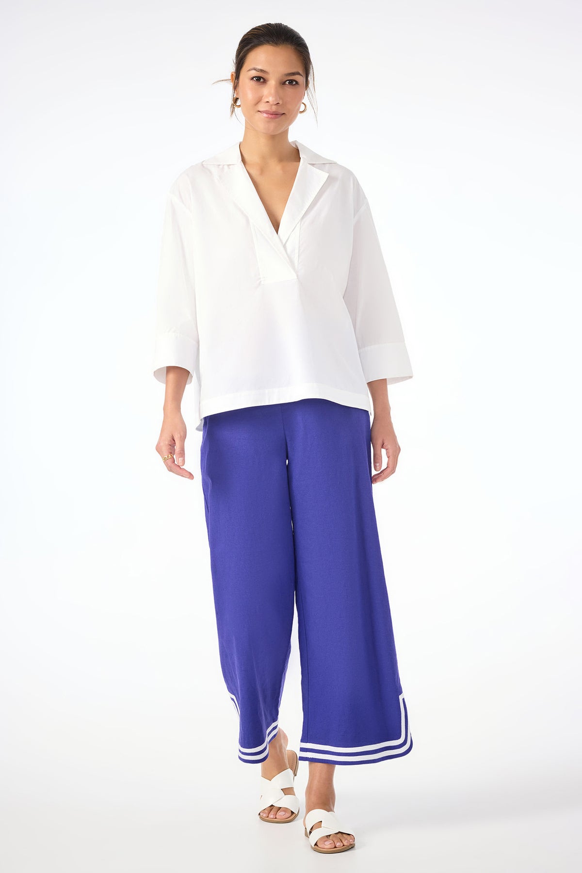 A woman stands against a white background wearing the Envie Top, wide-legged royal blue pants with white trim at the hem, and white slide sandals. Her hair is tied back and she smiles slightly.