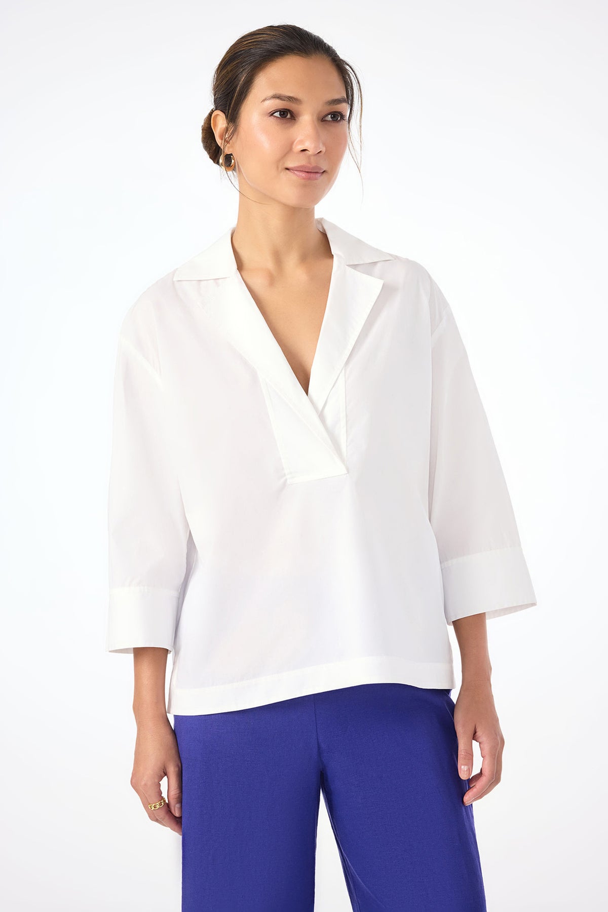 A woman poses against a plain white background, wearing the Envie Top in white—a loose-fitting blouse with three-quarter sleeves—paired with vibrant blue pants and looking slightly to the side with a relaxed expression.