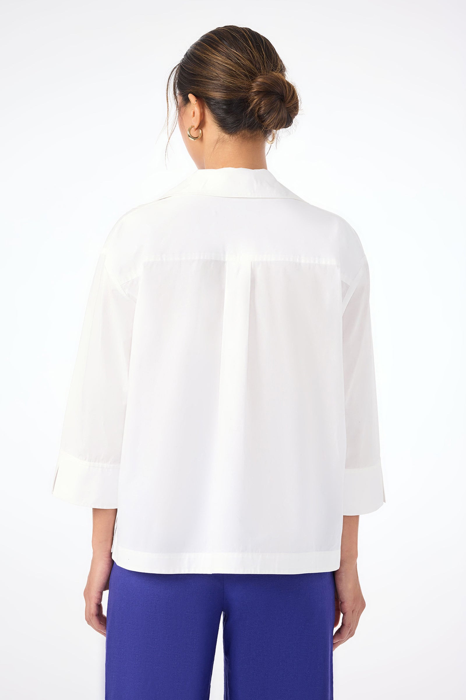 A woman with a neat low bun faces away, wearing the Envie Top—white, loose-fitting, with three-quarter sleeves—and bright blue pants against a plain white background.