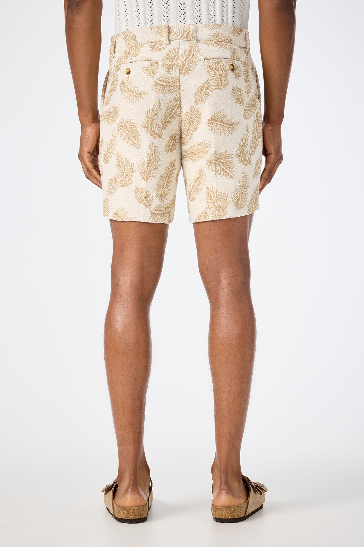 A person is seen from the back wearing the Lawrence Short - Desert Fronds Jacquard, featuring a brown leaf pattern, paired with a white textured shirt and tan slip-on shoes, standing against a plain white background.
