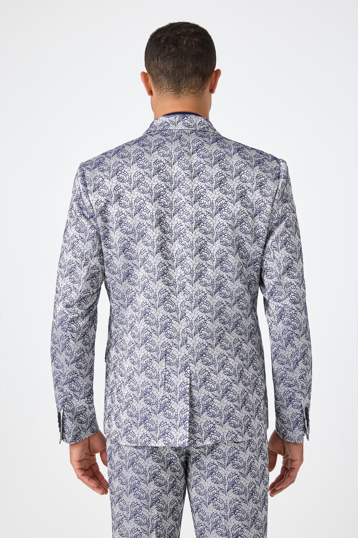 A man is seen from behind wearing the Thurston Blazer - Efflorescent Jacquard, a matching suit jacket and pants set with an ornate silver and blue leafy pattern, against a plain white background.