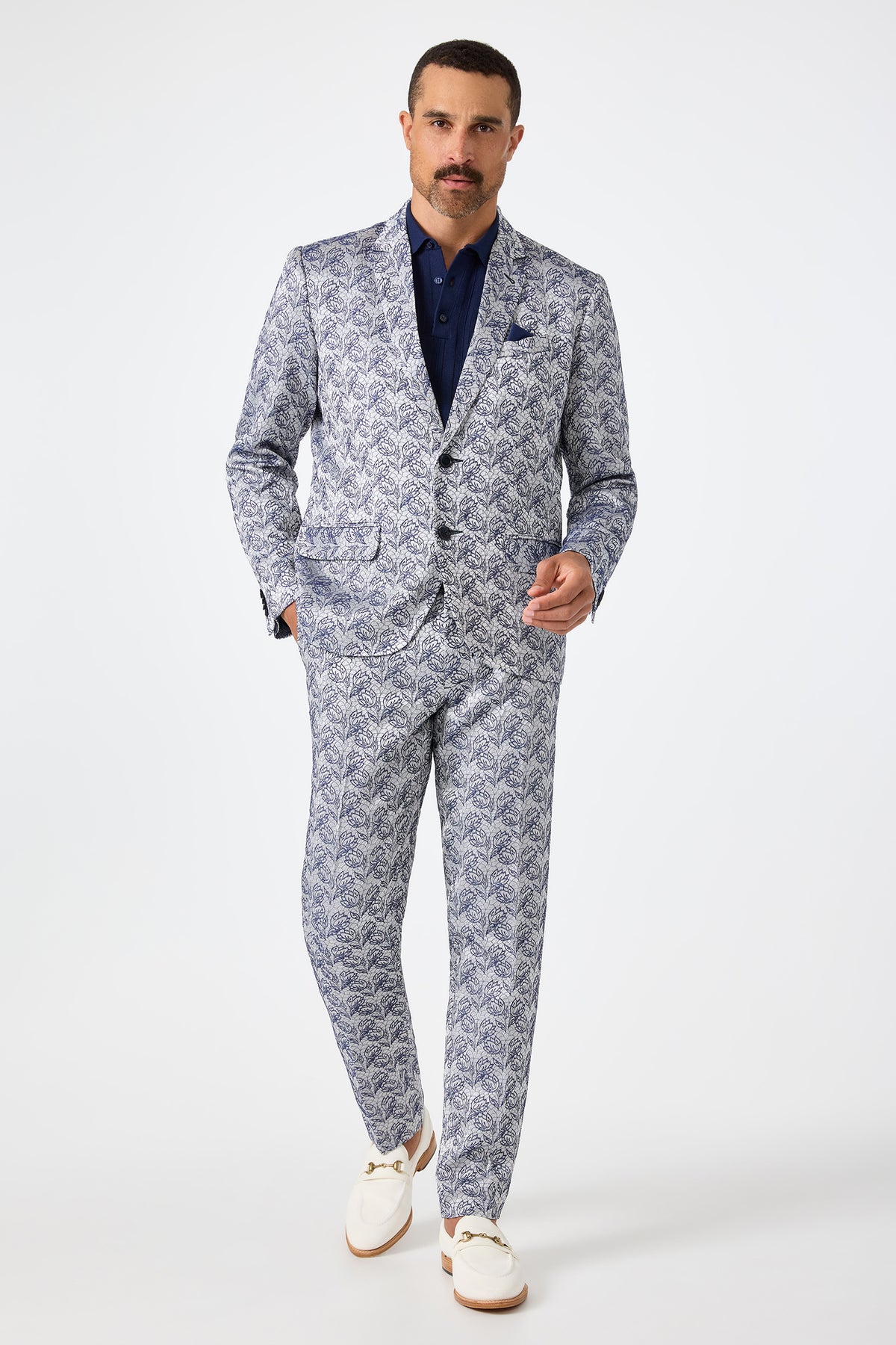 A man with a mustache stands confidently against a plain white background, dressed in the Clyde Slim Trouser - Efflorescent Jacquard, paired with a matching blue patterned suit, dark blue shirt, pocket square, and white loafers.