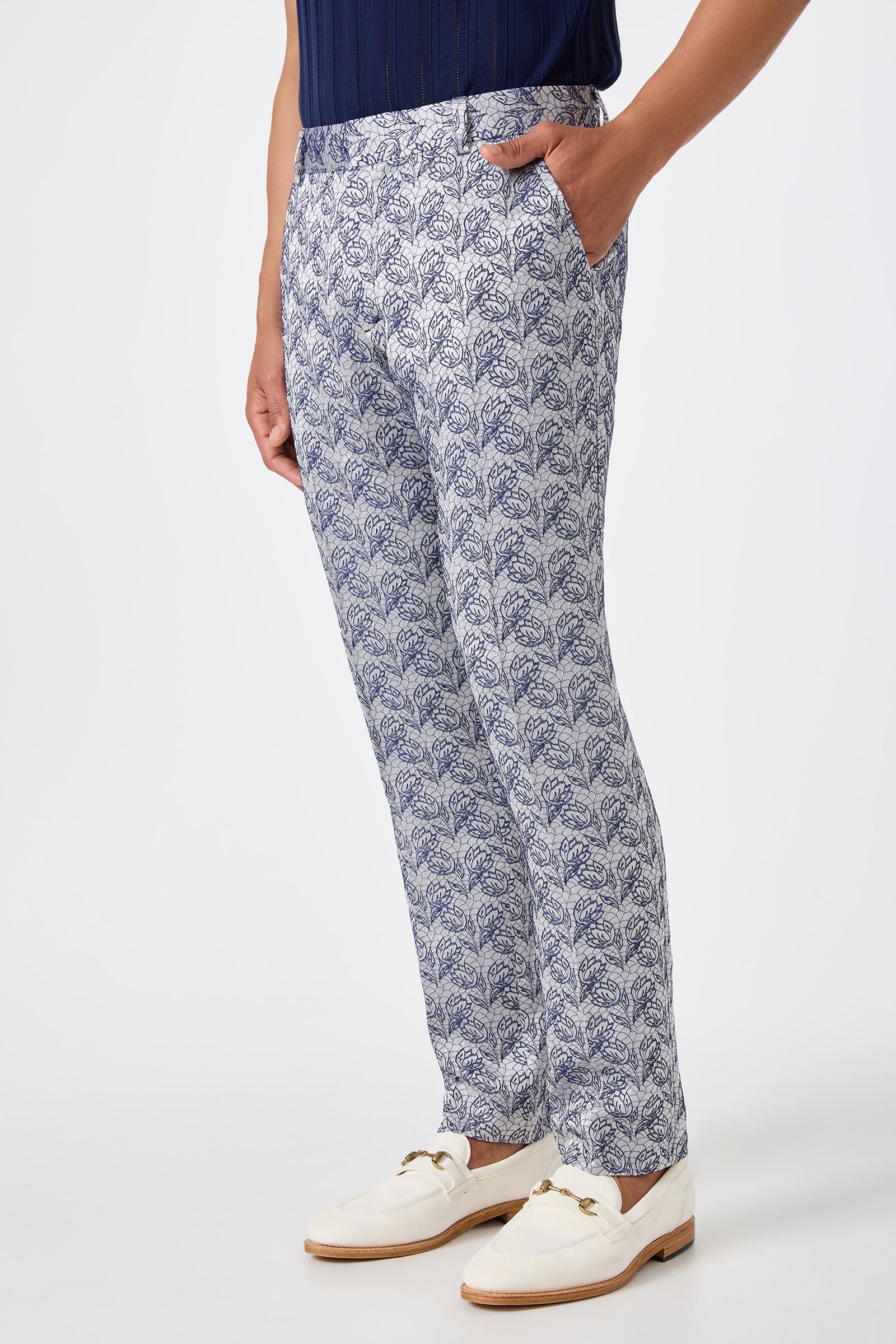 Model wears the Clyde Slim Trouser – Efflorescent Jacquard, featuring a patterned white and blue design, with a dark blue top and white loafers. Hands in pockets, standing against a plain white background.