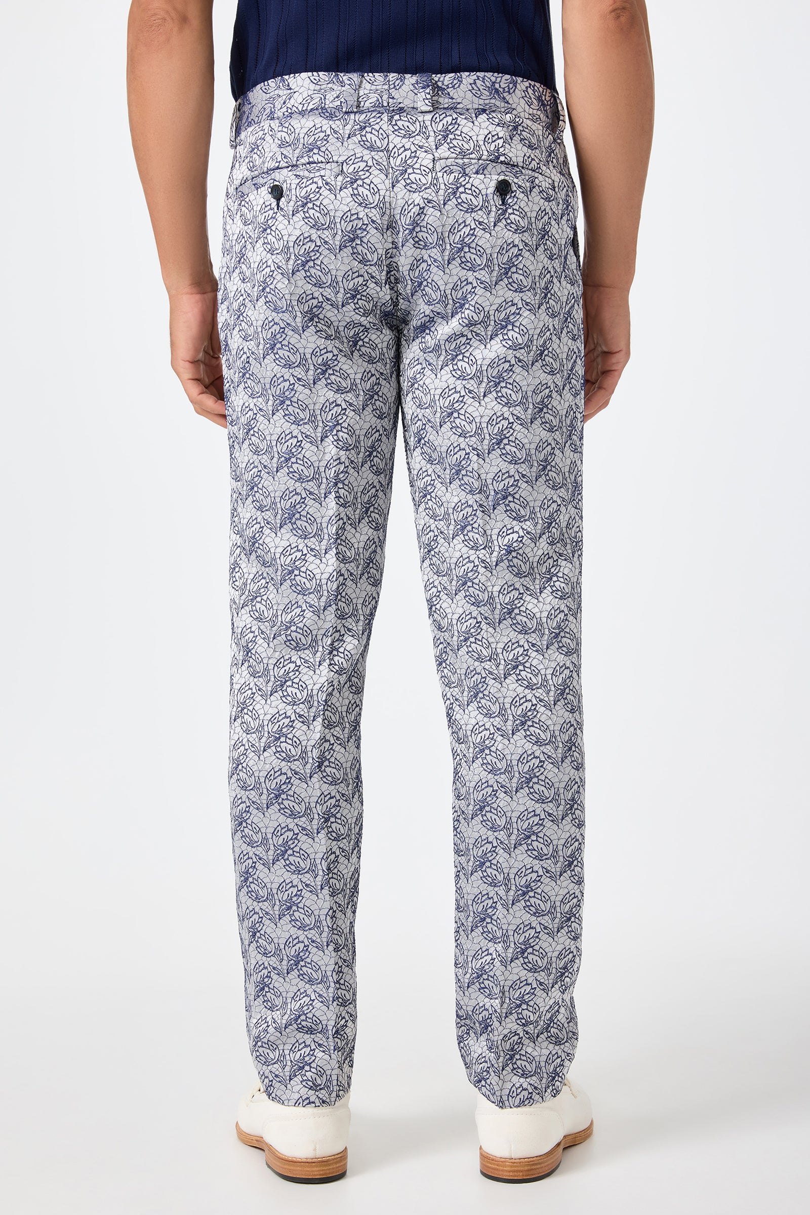 A person stands with their back to the camera, wearing the Clyde Slim Trouser - Efflorescent Jacquard, featuring a white base with a detailed navy leaf pattern, paired with a dark top and white shoes against a plain light background.