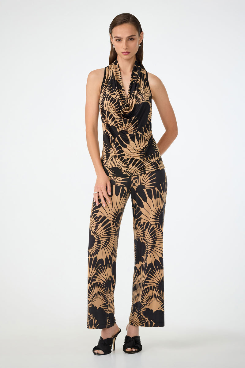 A woman with straight brown hair stands against a plain background wearing the Mosiac Cowl Neck Top in Juniper Jersey, featuring a sleeveless design and bold black and tan leaf pattern, paired with black open-toe heels.