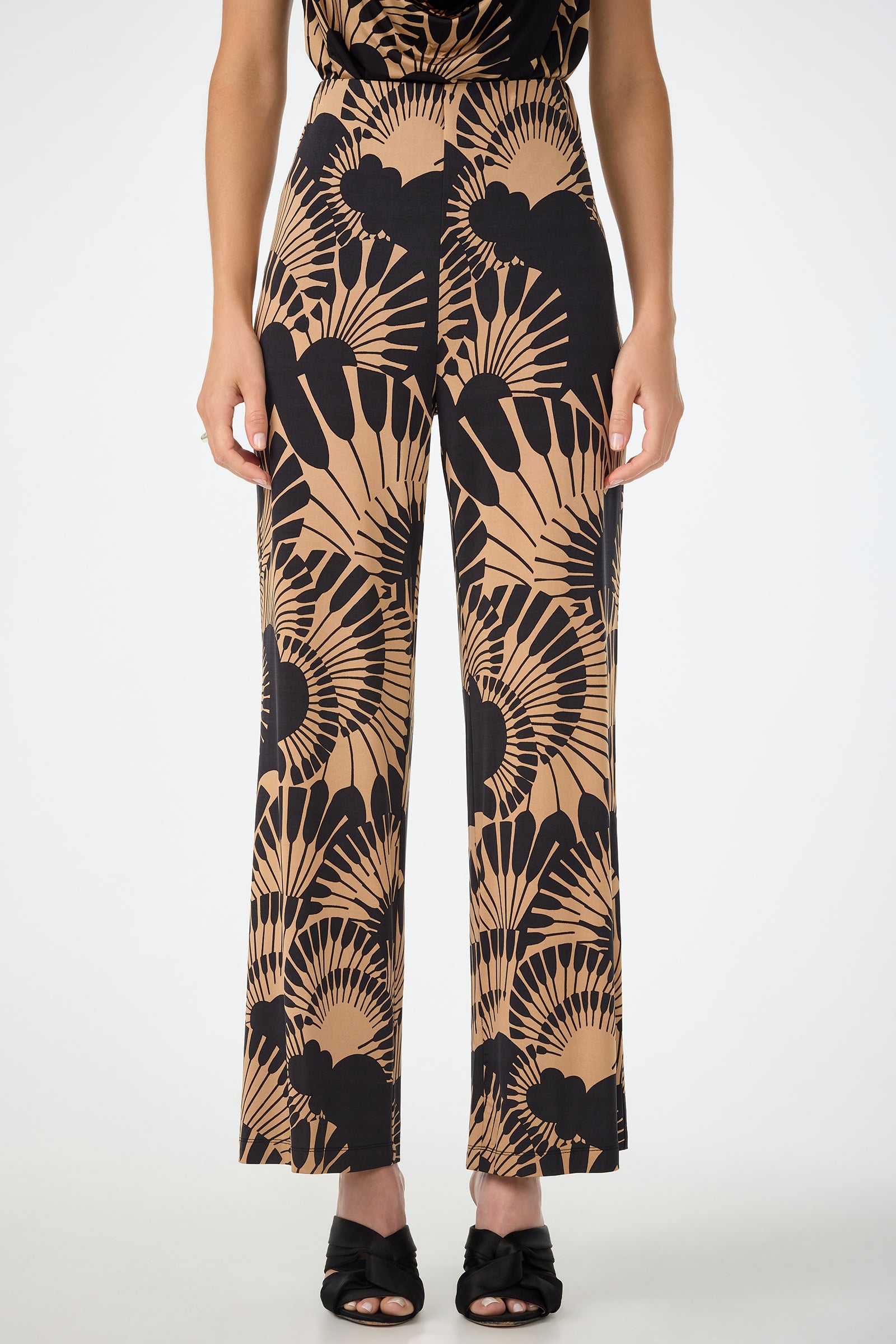 Someone wears Atoll Wide-Leg Cropped Pants in Juniper Jersey featuring a bold black and tan abstract fan pattern, paired with black open-toe heels, standing against a plain white background.