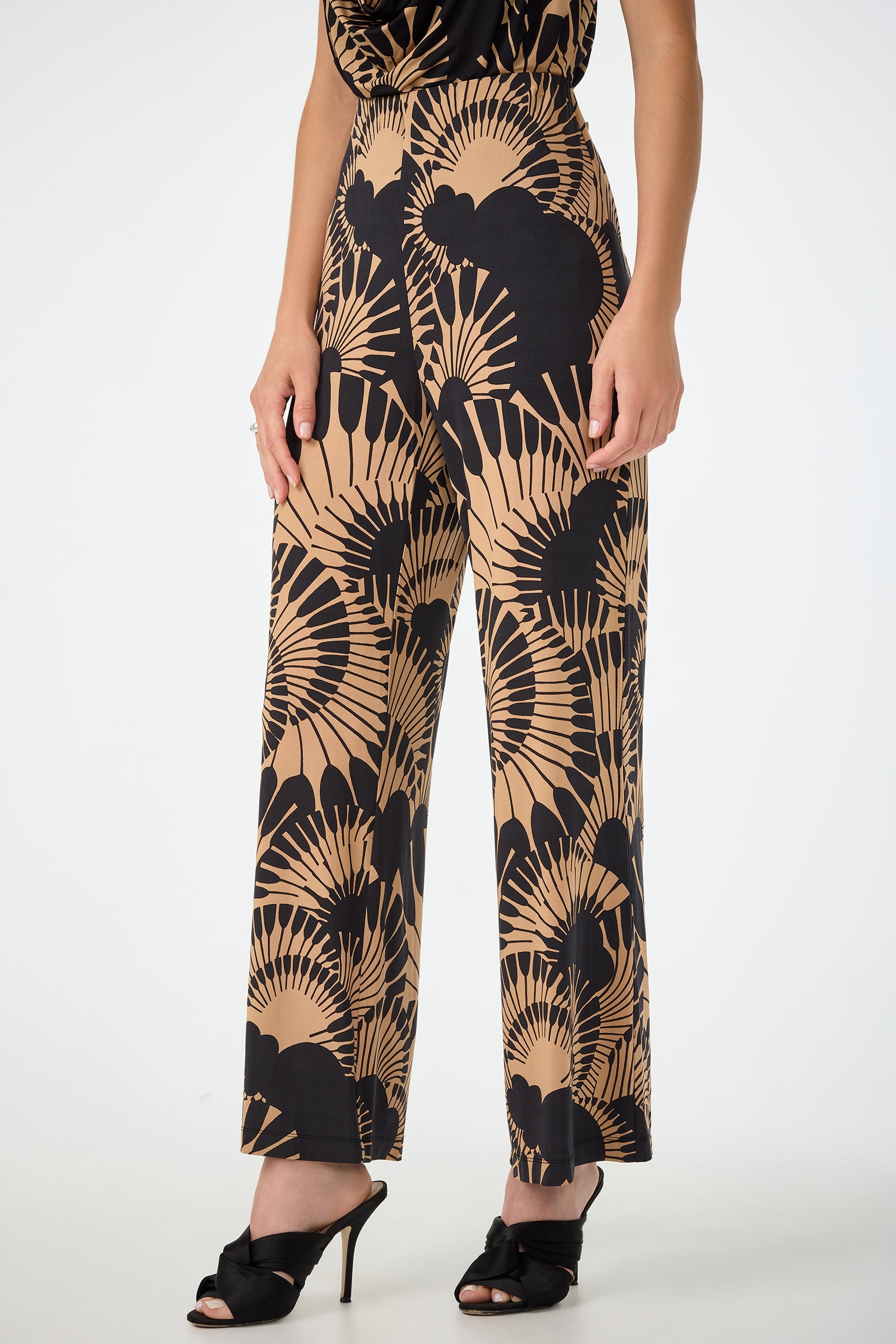 A woman wears the Atoll Wide-Leg Cropped Pants - Juniper Jersey, featuring a tan and black abstract fan pattern, paired with a black top and black open-toe heeled shoes. Only her lower body is shown.