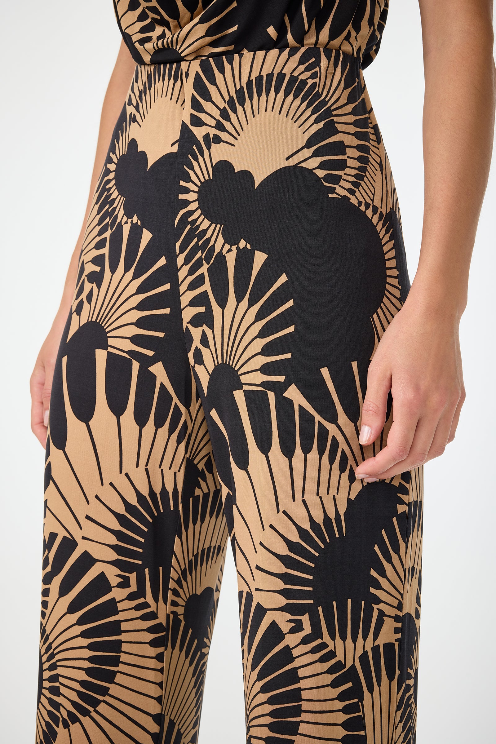 A person wears the Atoll Wide-Leg Cropped Pants in Juniper Jersey, featuring a bold black abstract pattern with fan and cloud motifs on beige fabric. Only their lower torso and upper legs are visible, one arm relaxed at their side.