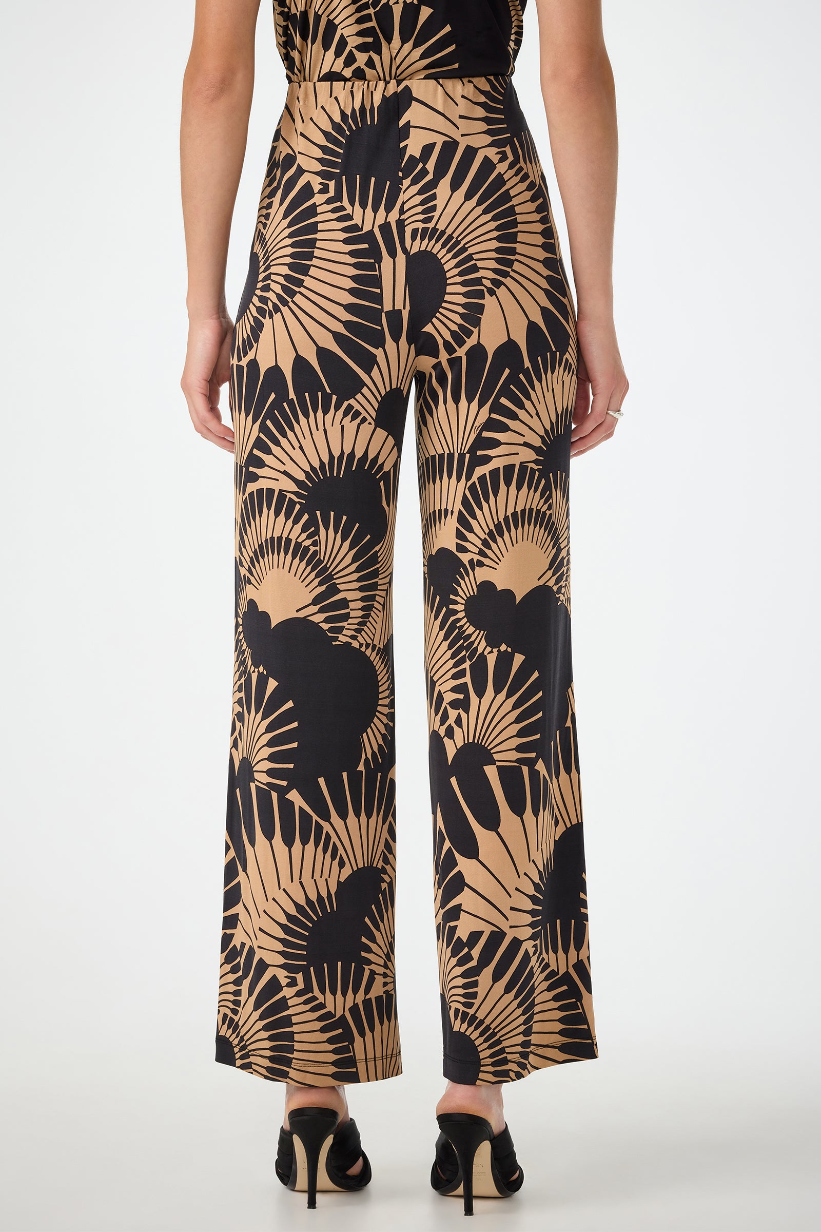 A person stands facing away, wearing Atoll Wide-Leg Cropped Pants in Juniper Jersey—a high-waisted style with a tan and black abstract fan pattern—paired with black heeled sandals against a plain white background.