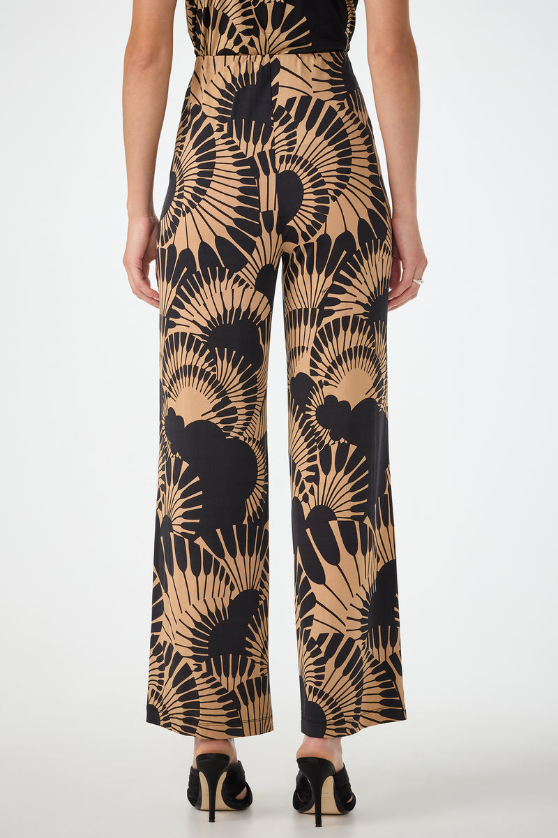 A person stands facing away, wearing Atoll Wide-Leg Cropped Pants in Juniper Jersey—a high-waisted style with a tan and black abstract fan pattern—paired with black heeled sandals against a plain white background.