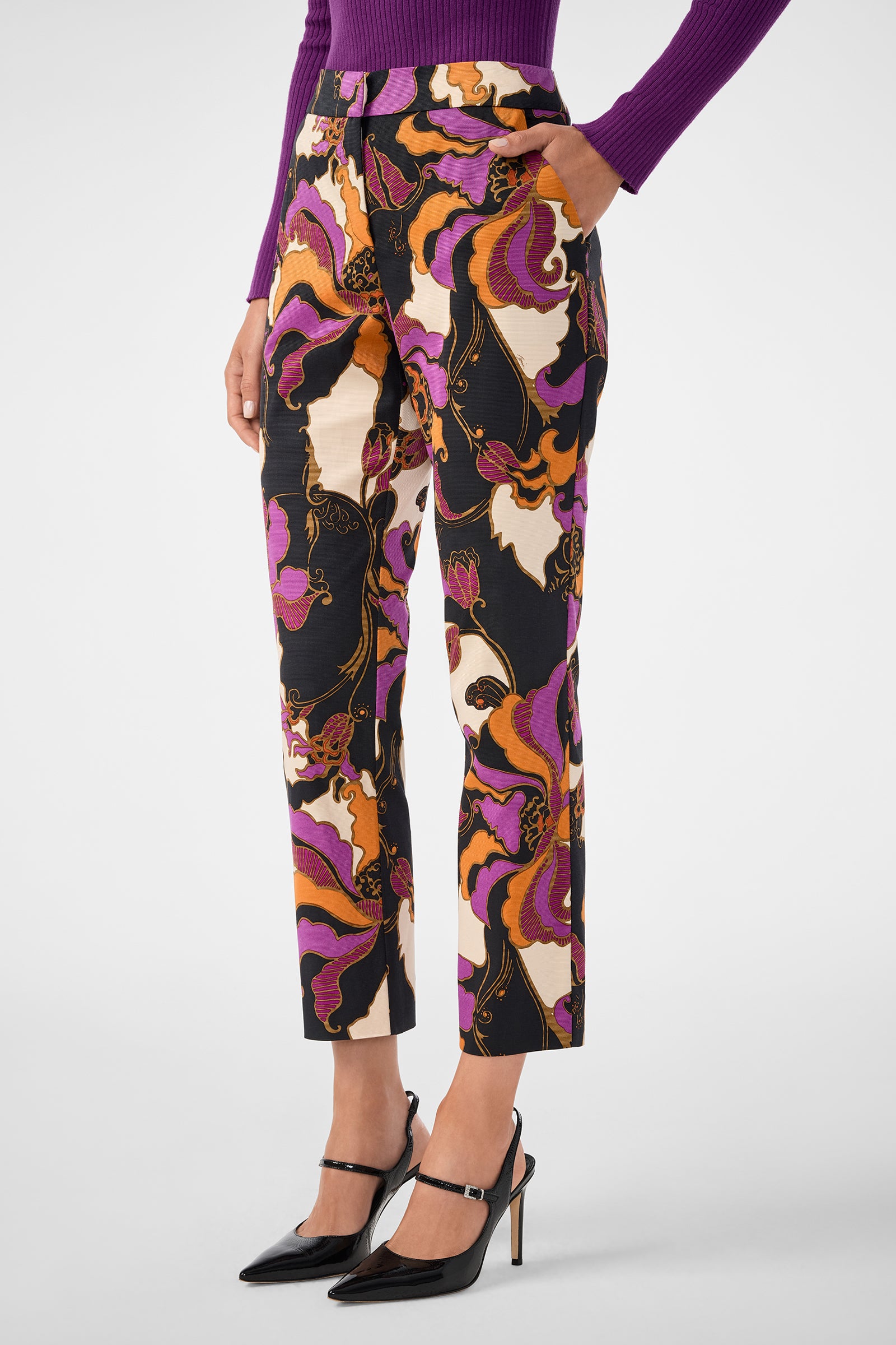 Someone wears the Moss 2 Pant—high-waisted, ankle-length pants featuring a bold abstract print in purple, orange, black, and white—paired with a purple long-sleeve top and black pointed high heels.