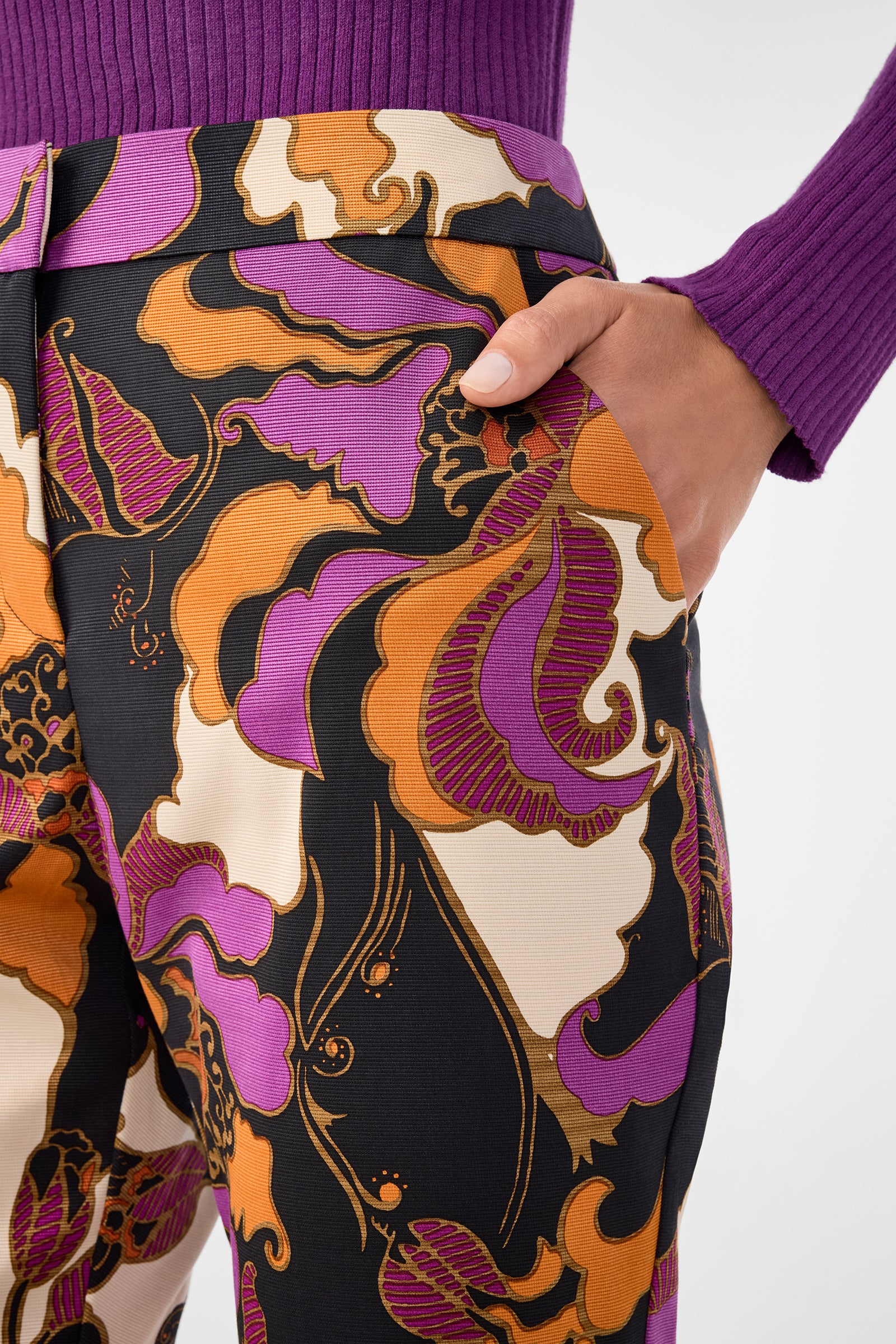 Someone wearing the Moss 2 Pant, which features orange, purple, and cream floral patterns, rests a hand in the pocket. They pair the pants with a ribbed, long-sleeve purple top.