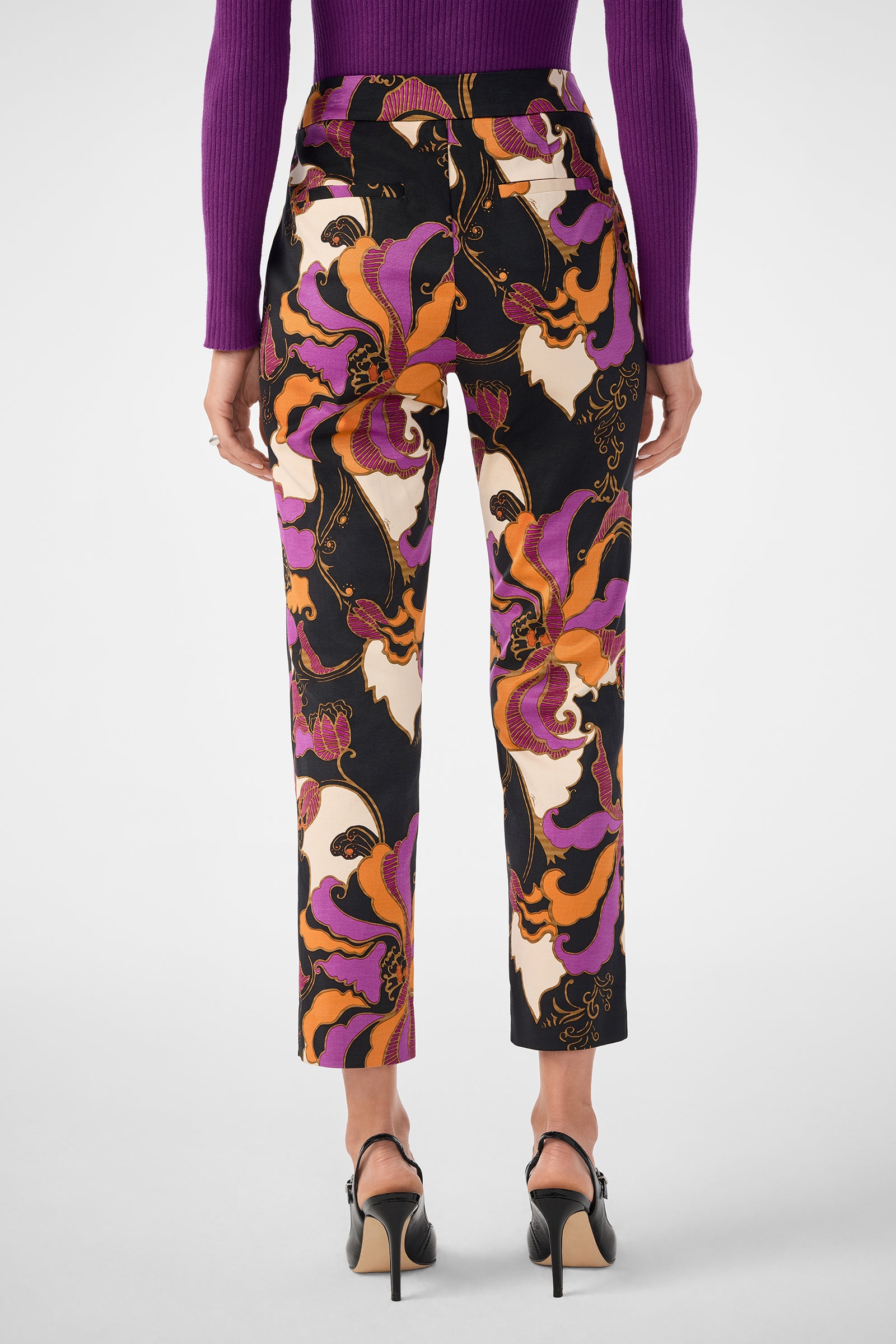 A person shown from the back wears the Moss 2 Pant—high-waisted, ankle-length with a bold orange, purple, black, and white abstract floral print—paired with a purple ribbed long-sleeve top and black heeled sandals.