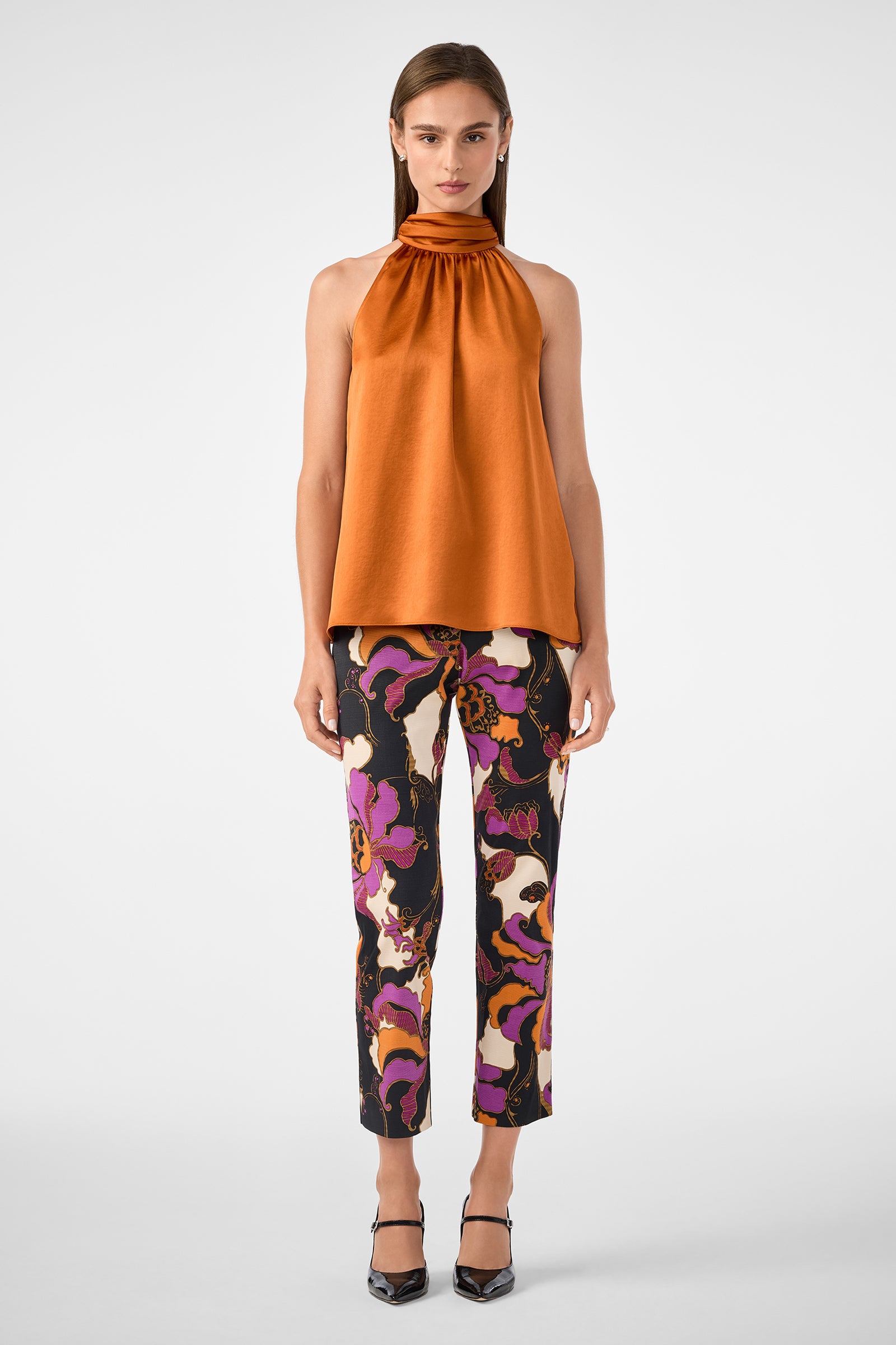 A woman faces forward in a sleeveless orange high-neck top and Moss 2 Pant featuring a colorful abstract pattern in purple, yellow, and black, paired with black heels against a plain white background.