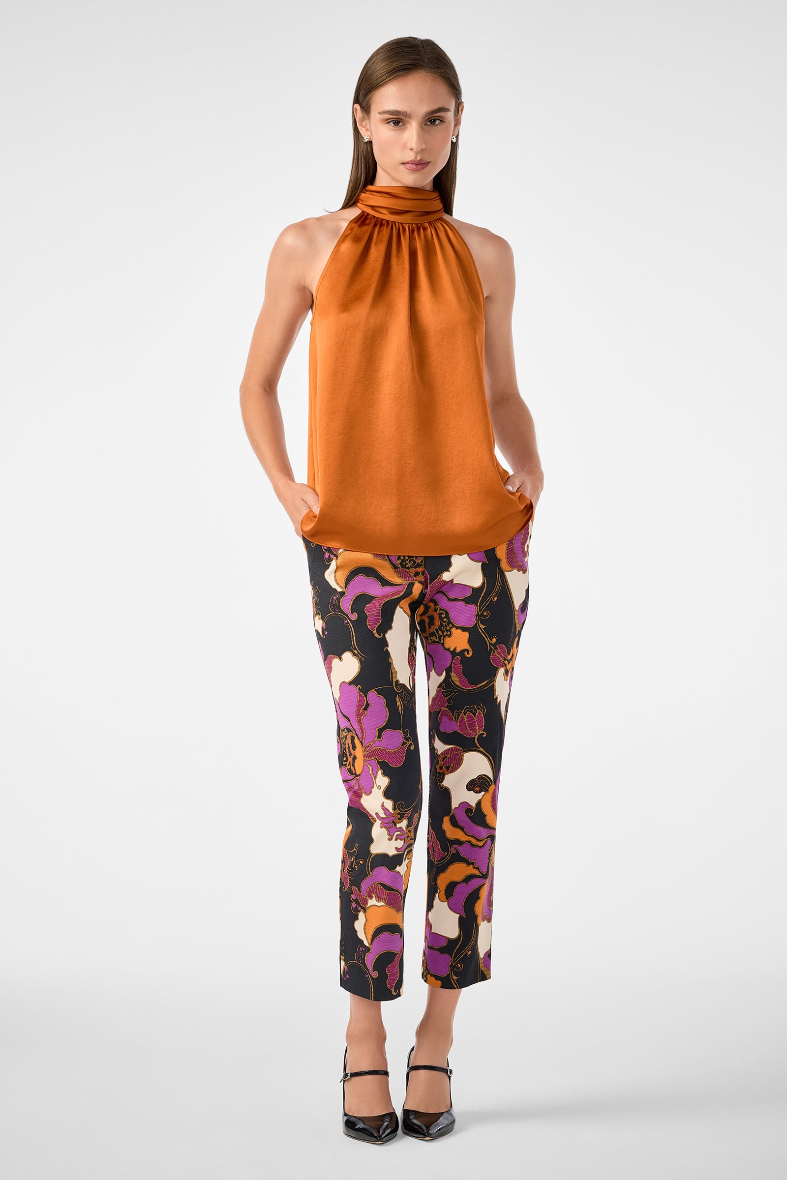 A woman wears the Zana Top, a sleeveless high-neck orange shirt, paired with colorful abstract-patterned pants in purple, black, and pink shades and black heels, standing against a plain light background.