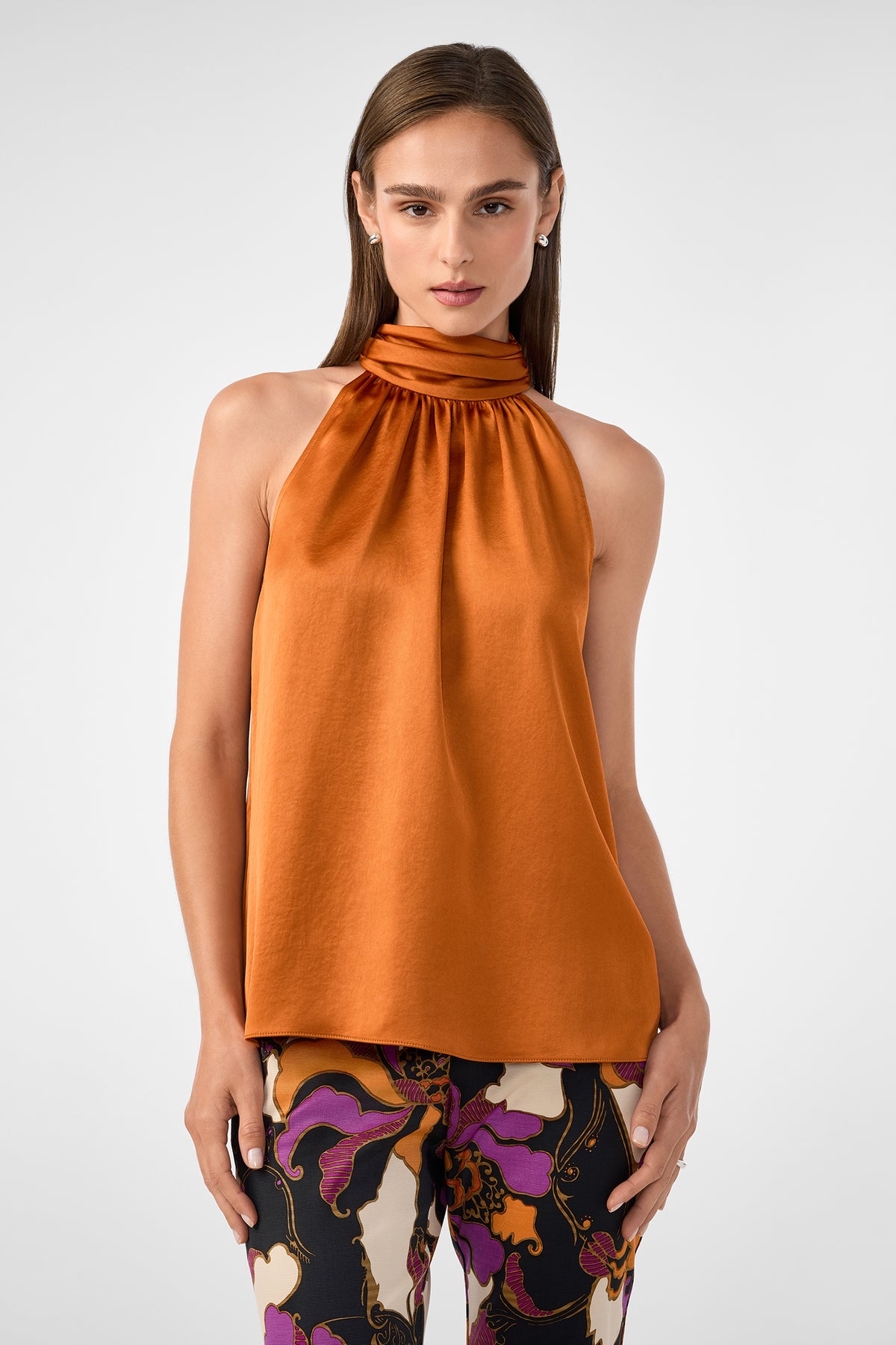 A woman wears the Zana Top, a sleeveless high-neck satin blouse in burnt orange, paired with colorful floral pants, standing against a plain light background.
