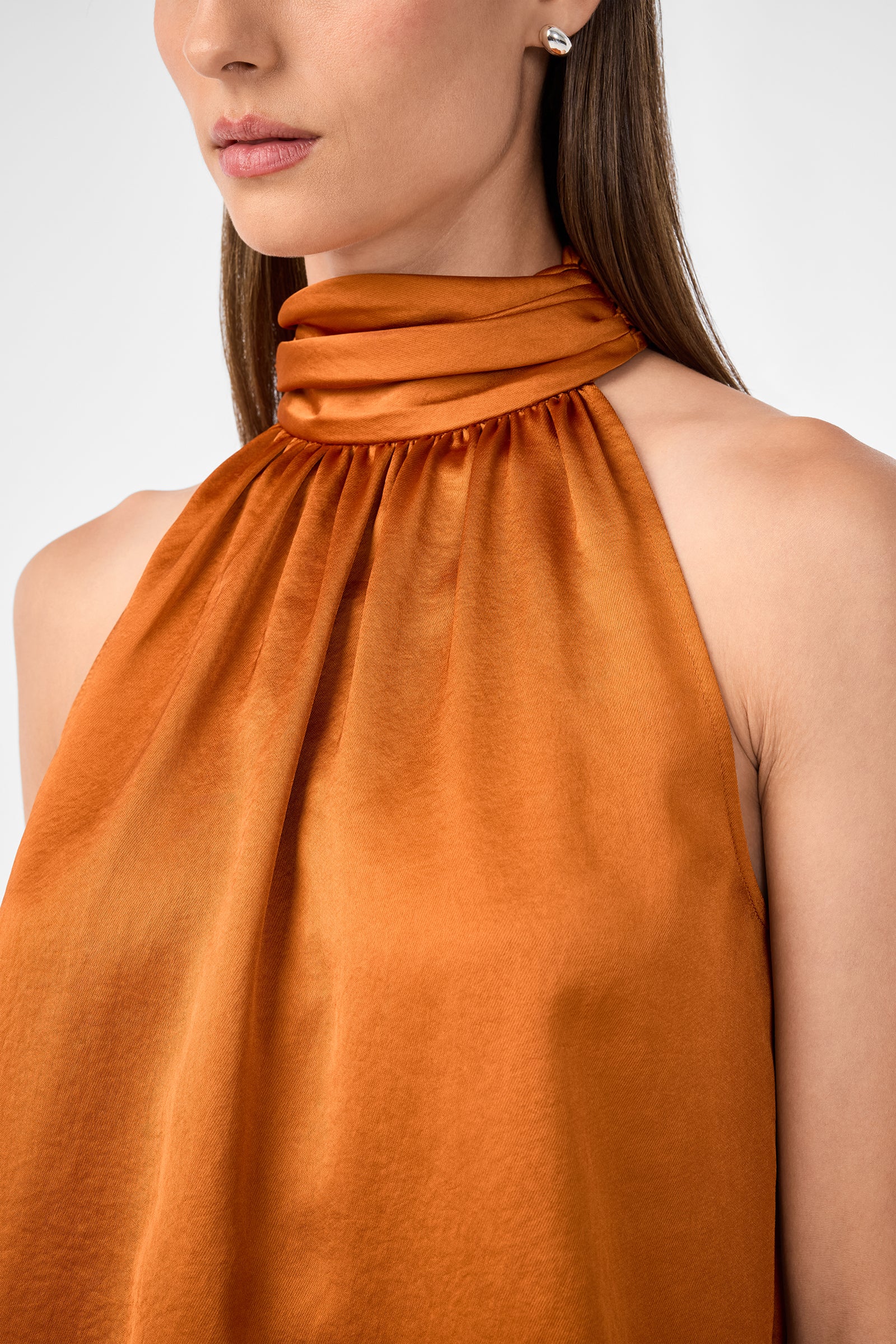 A woman wears the Zana Top—a sleeveless, high-neck satin blouse in rust with pleated detailing at the neckline. She has straight brown hair, pearl stud earrings, and is posed against a neutral background.