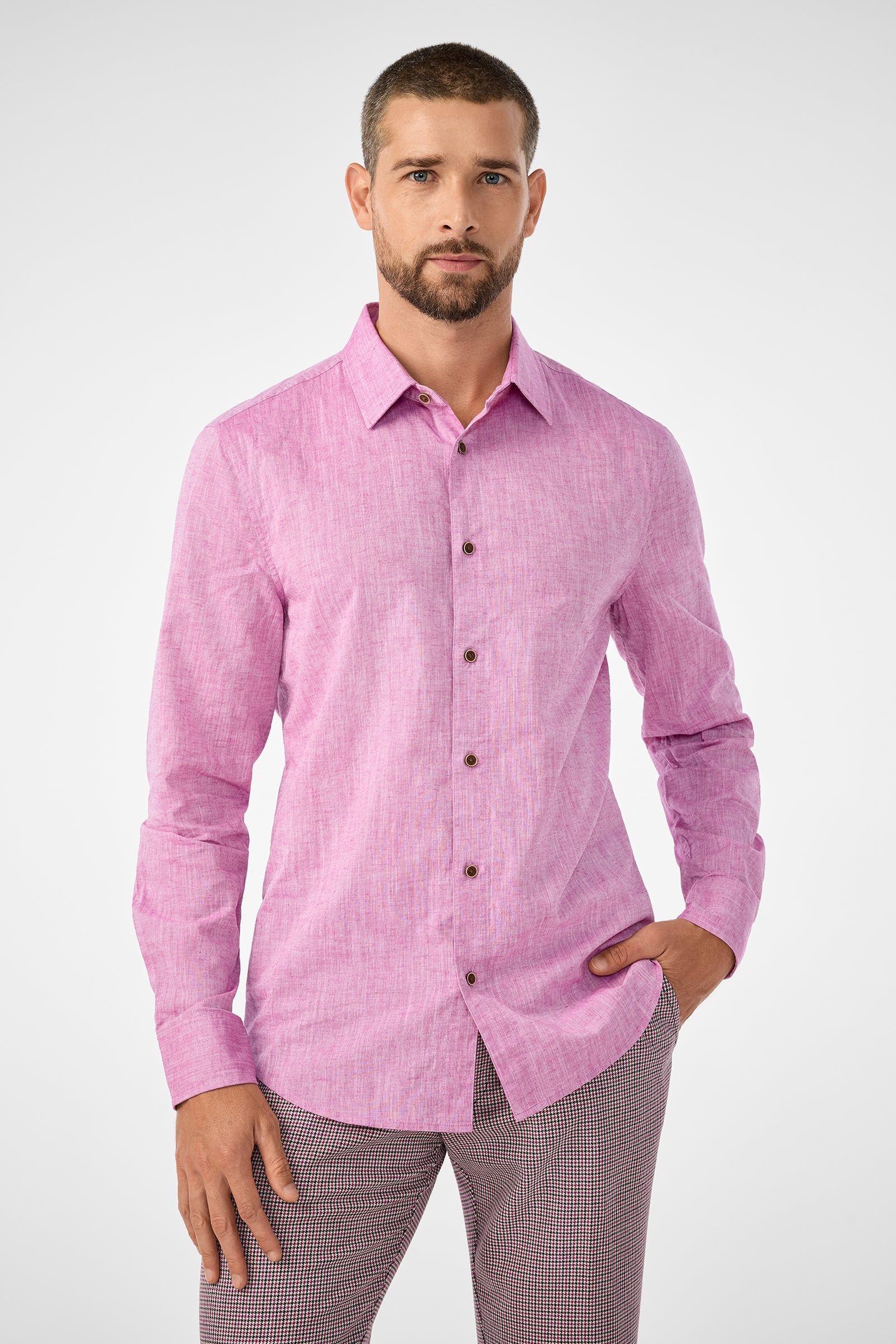 A man with short hair and a beard wears the Aaron Slim Fit Long-Sleeve Shirt in Geranium and checkered pants, standing confidently with one hand in his pocket against a plain, light background.