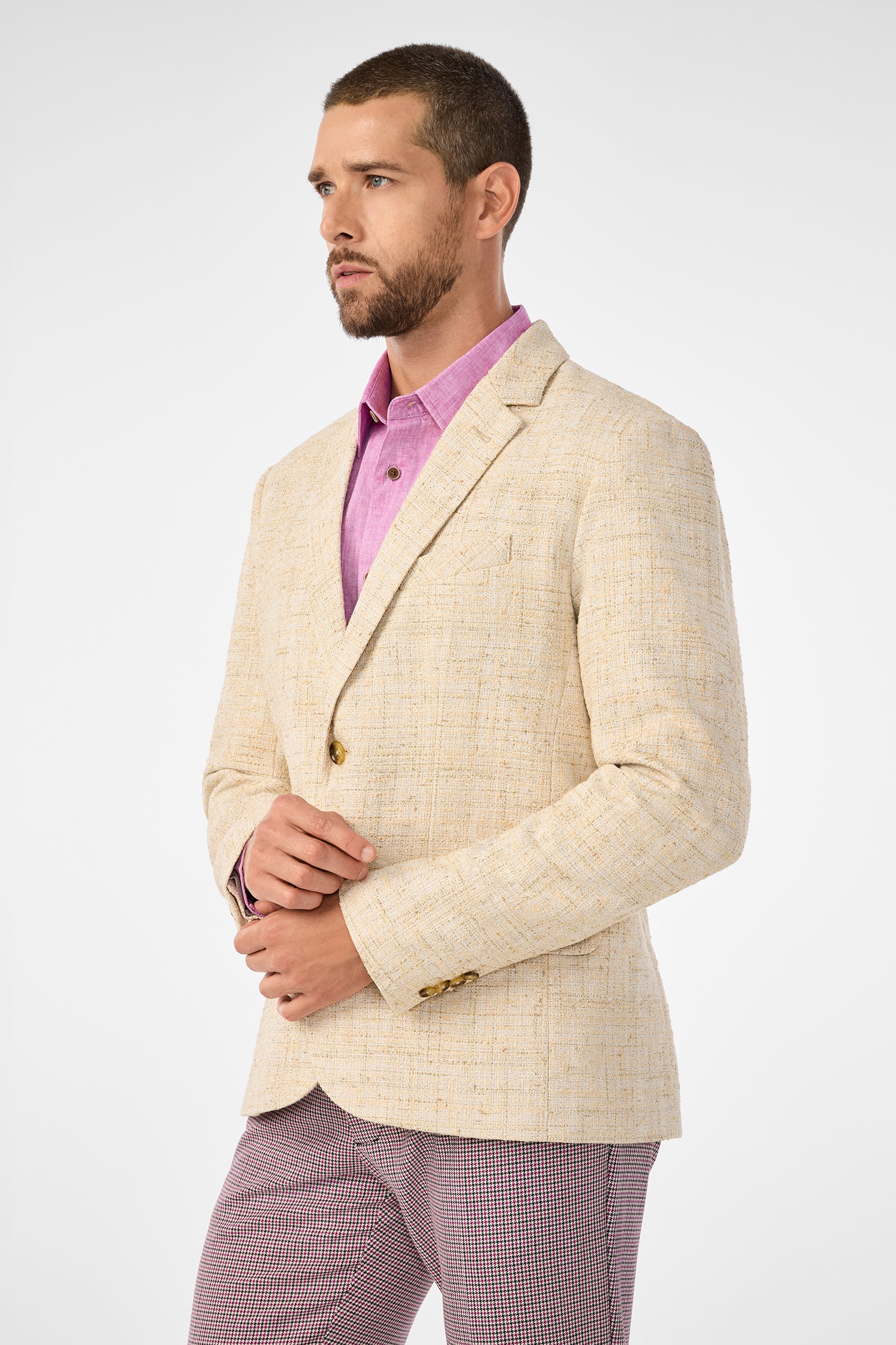 A man with short hair and a trimmed beard models the Thurston Blazer in beige over a pink dress shirt and patterned pants, standing against a plain white background.