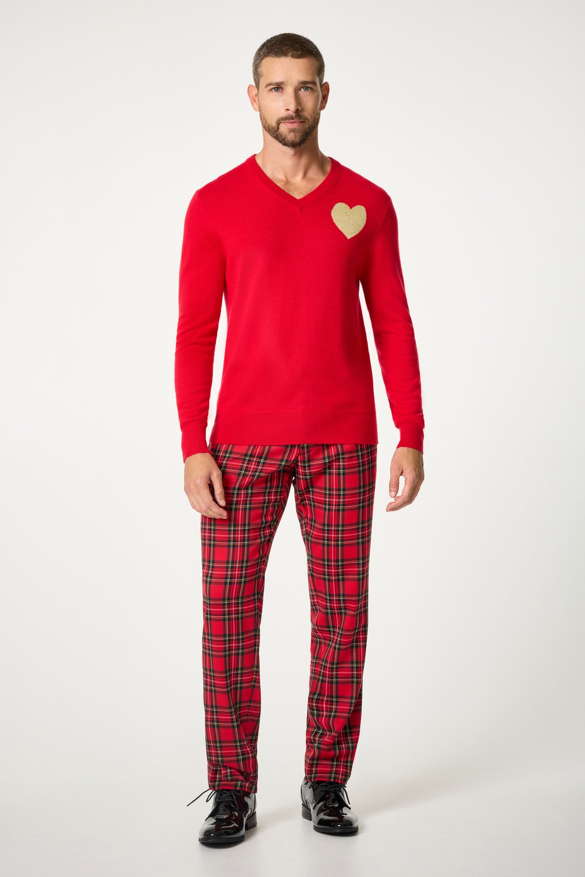 A man with short hair and a beard stands against a plain background wearing the Lover V-Neck Sweater, featuring a gold heart on the chest, red plaid pajama pants, and black shoes.