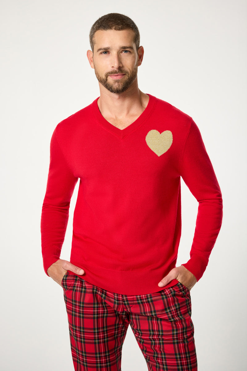 A man with short hair and a beard wears the Lover V-Neck Sweater, a bright red top featuring a gold heart on the chest, paired with red plaid pants. He stands with his hands in his pockets against a simple light background.