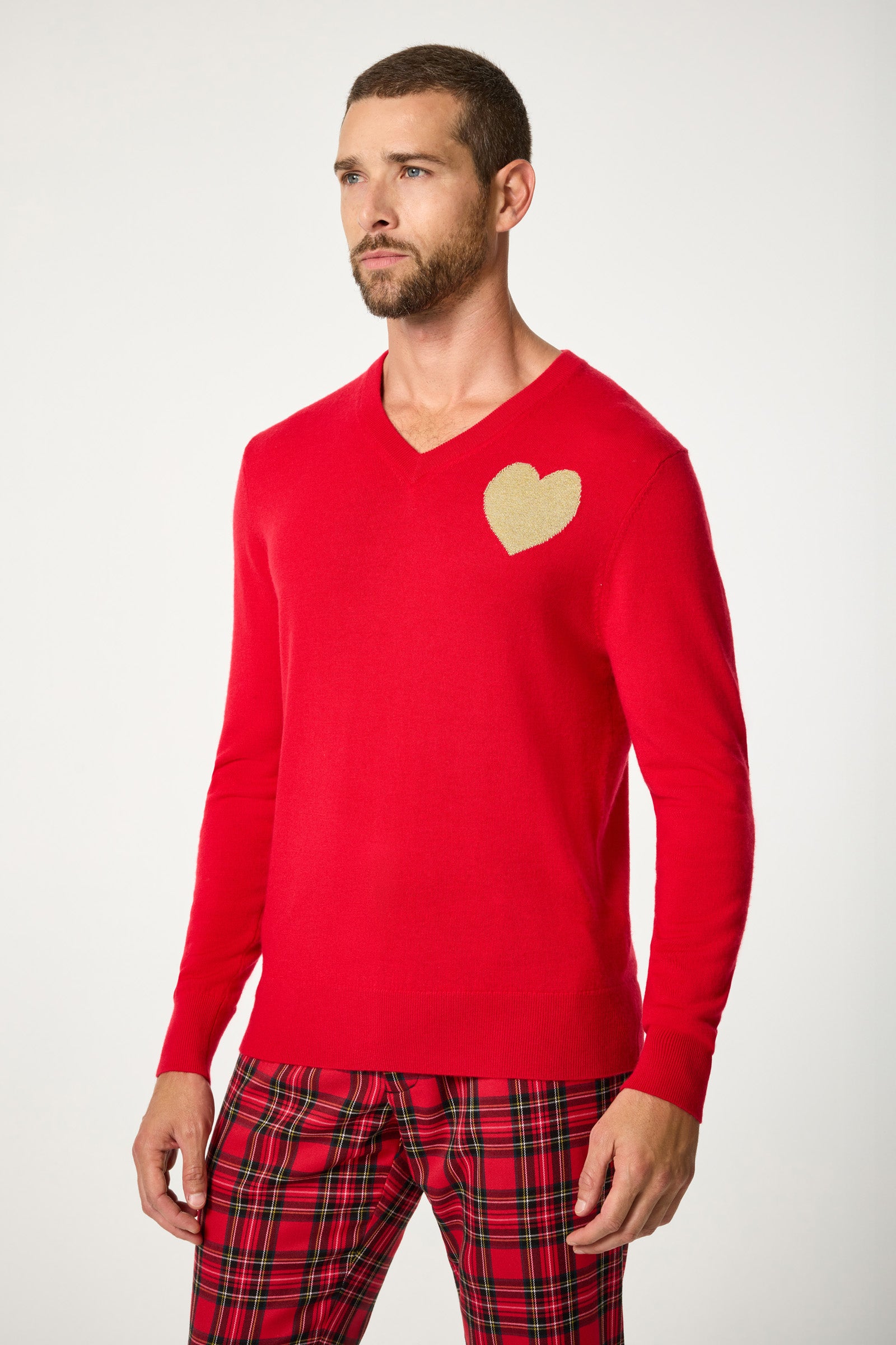 A man wears the Lover V-Neck Sweater, featuring a bright red color and a gold heart on the chest, paired with red plaid pants, standing against a plain white background.