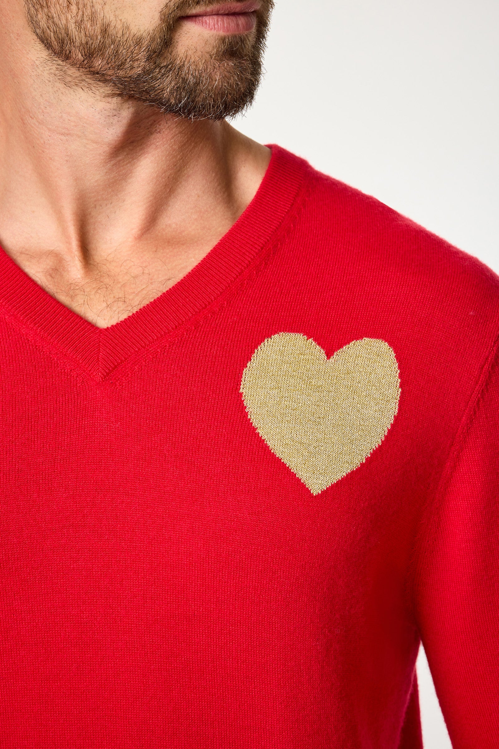 Someone wearing the Lover V-Neck Sweater, a bright red sweater with a large gold heart design on the left chest. Only the lower part of their face and neck are shown.