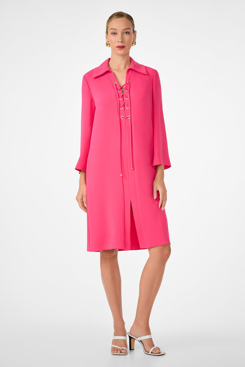 A woman stands against a white background wearing the Janeiro Dress, a bright pink, knee-length style with a lace-up front and three-quarter sleeves. She pairs it with white heeled sandals, gold earrings, and has her hair pulled back.