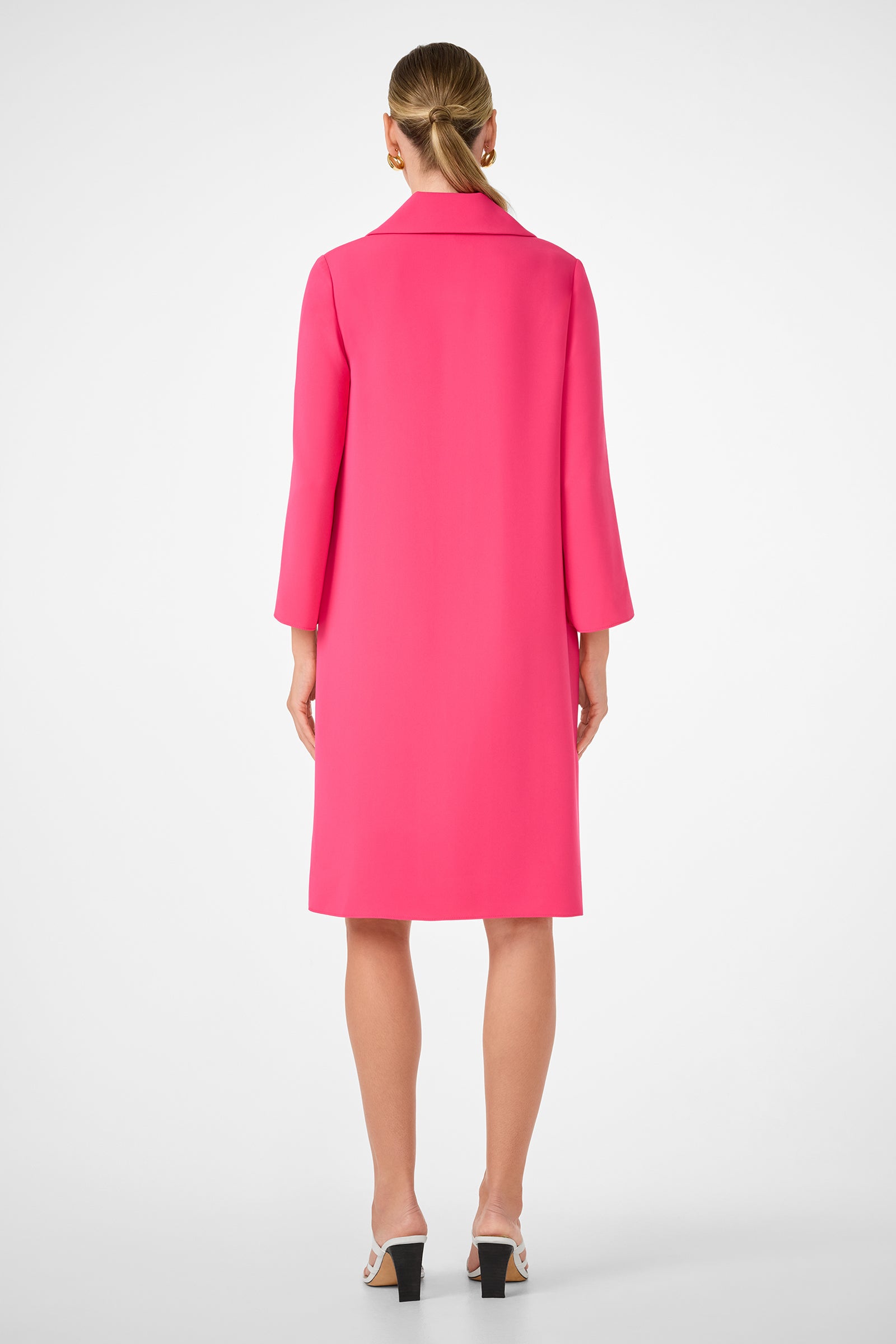 A woman faces away against a white background, wearing the Janeiro Dress in bright pink with three-quarter sleeves and knee-length hem, paired with white heeled sandals. Her hair is tied back in a low ponytail.