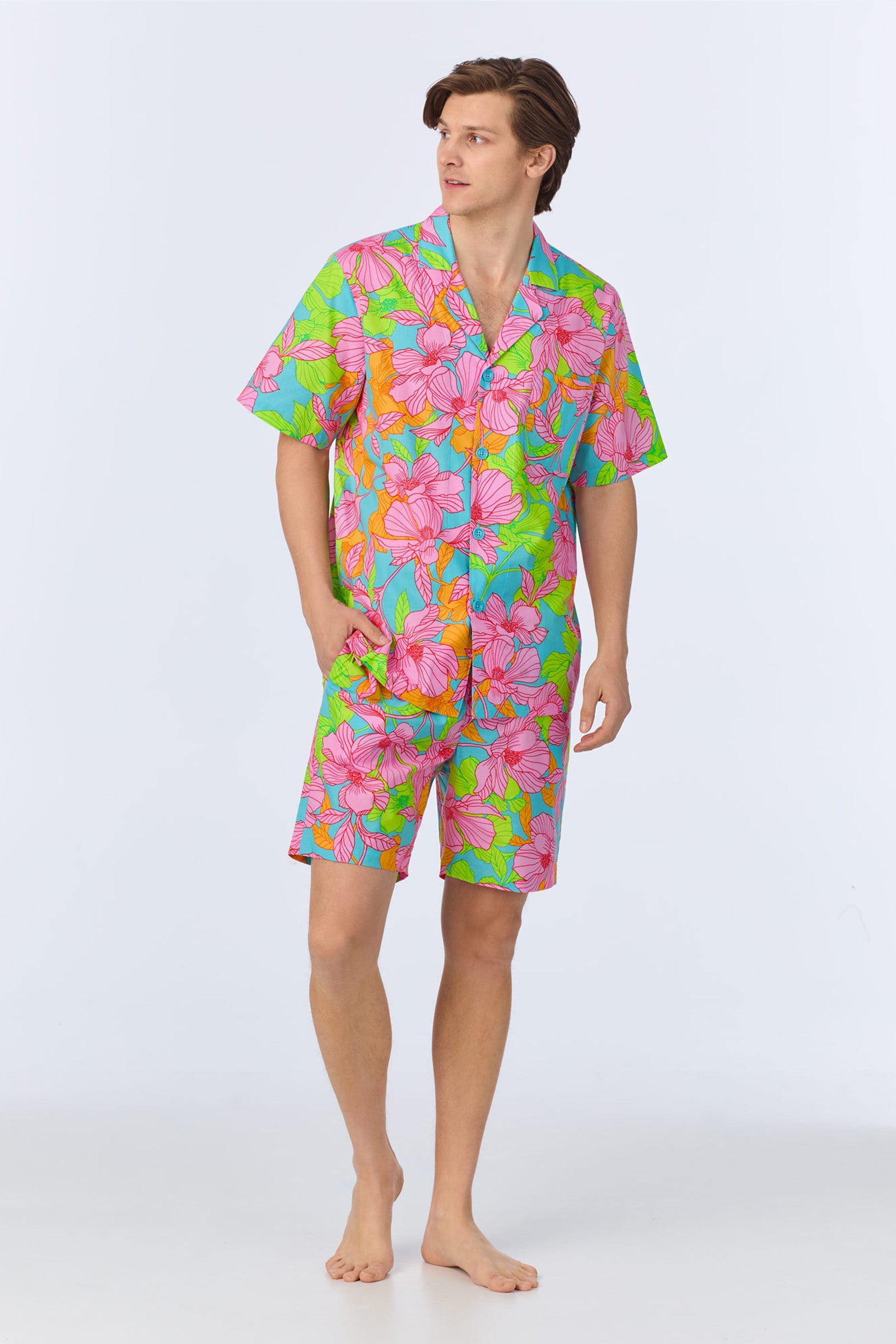 A man stands barefoot in the Mens Boxer PJ Set - Linear Floral Poplin, Multi. The short-sleeve button-up shirt and shorts feature a colorful linear floral pattern. He has one hand in his pocket and looks to the side.