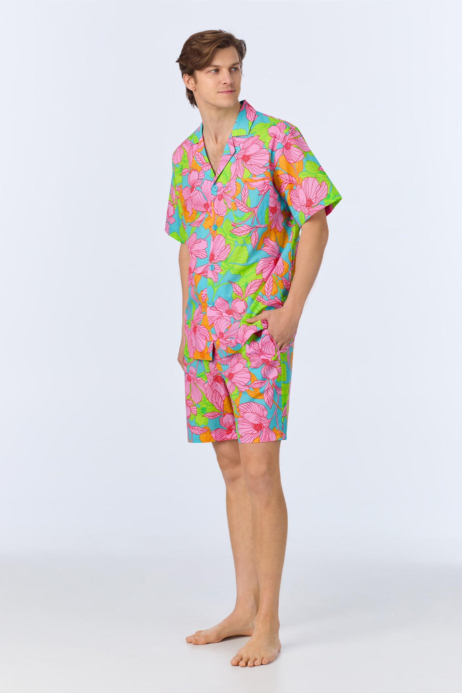 A man stands barefoot wearing the Mens Boxer PJ Set - Linear Floral Poplin, featuring a colorful short-sleeved shirt and matching shorts with a bright pink, green, and blue floral pattern against a plain light background.