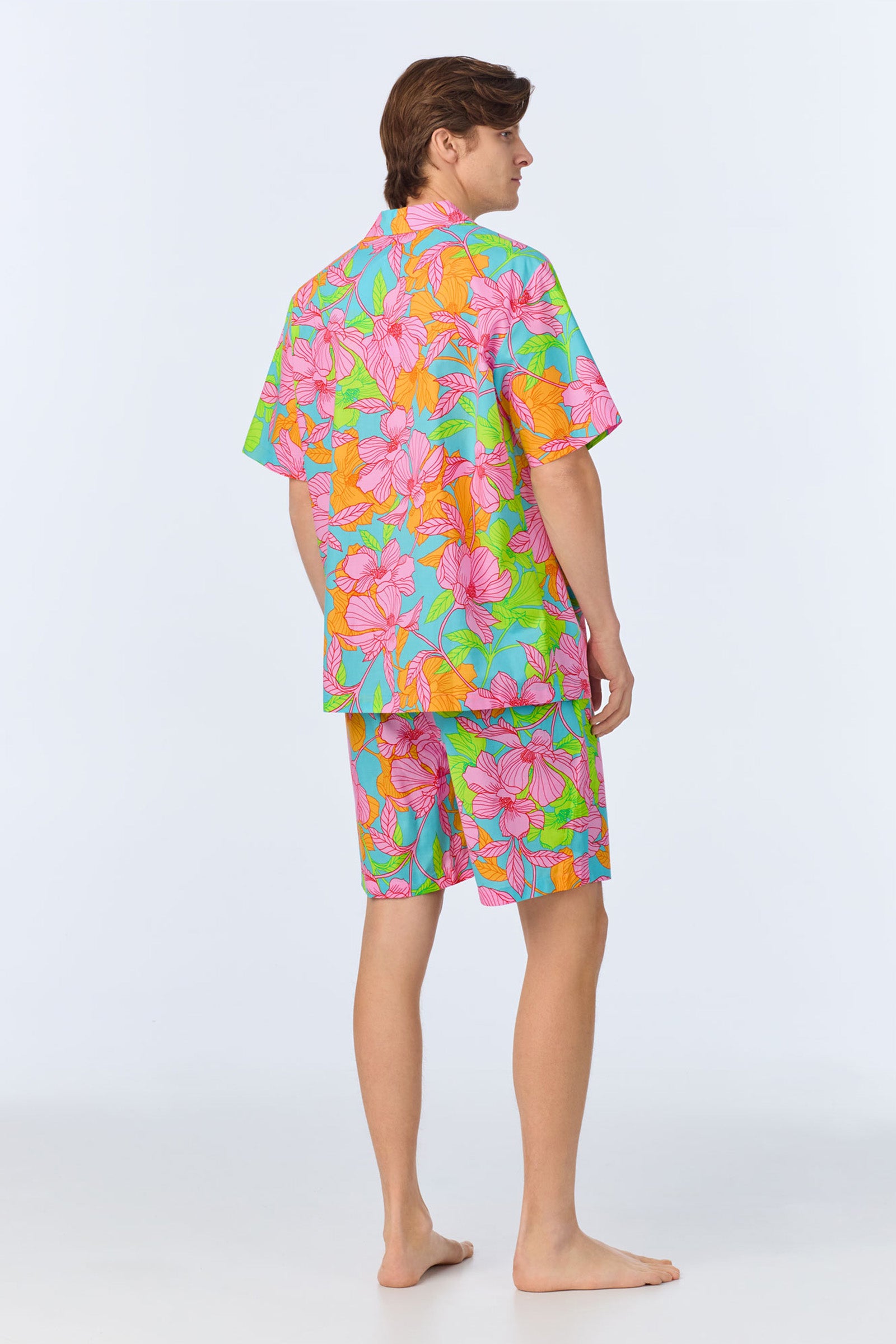 A person stands barefoot, facing away, wearing the Mens Boxer PJ Set - Linear Floral Poplin, a matching short-sleeve shirt and shorts set with a bright, multicolor floral pattern on a light background.