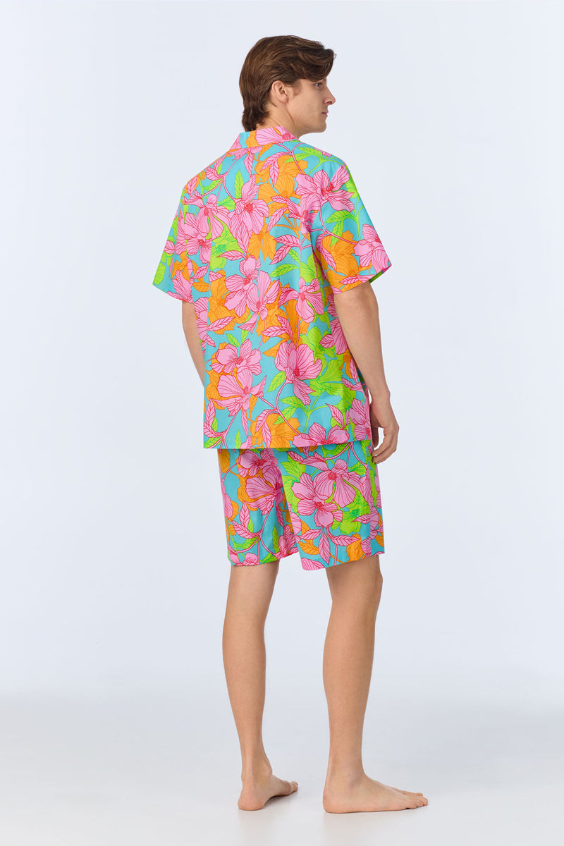 A person stands barefoot, facing away, wearing the Mens Boxer PJ Set - Linear Floral Poplin, a matching short-sleeve shirt and shorts set with a bright, multicolor floral pattern on a light background.